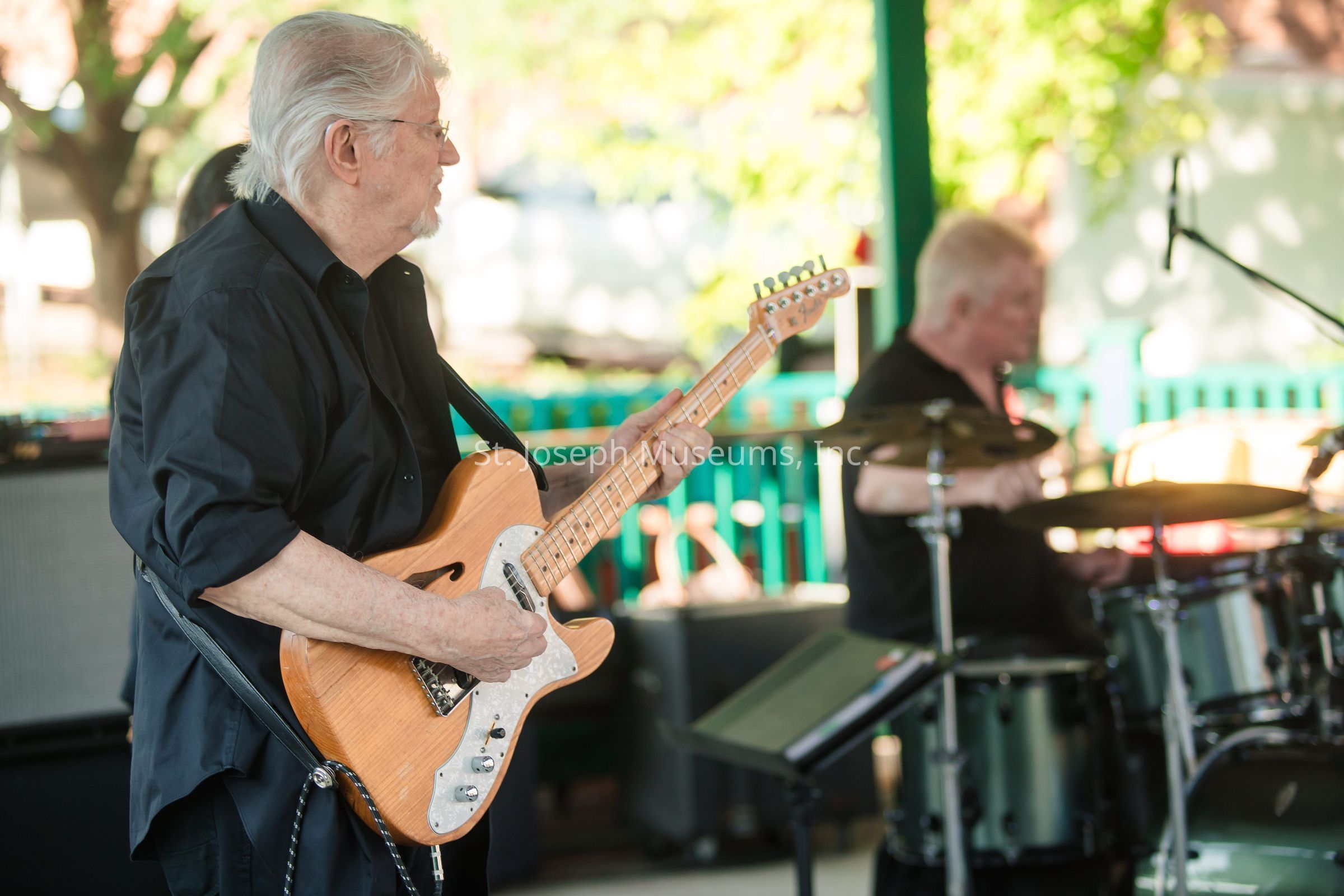 In the image, an older man with white hair and glasses is passionately playing an electric guitar while wearing a black shirt, standing under a shaded area, possibly a pavilion or gazebo, during a sunny day. He is deeply focused on his music, indicating experience and dedication, and is accompanied by another musician in the background who is playing the drums. The scene is set in an outdoor setting with blurred green foliage and a wooden fence, suggesting a casual live performance, potentially a community event or a casual gig. The guitarist's poise and concentration, coupled with the harmonious collaboration of the drummer, create a dynamic and engaging musical atmosphere.