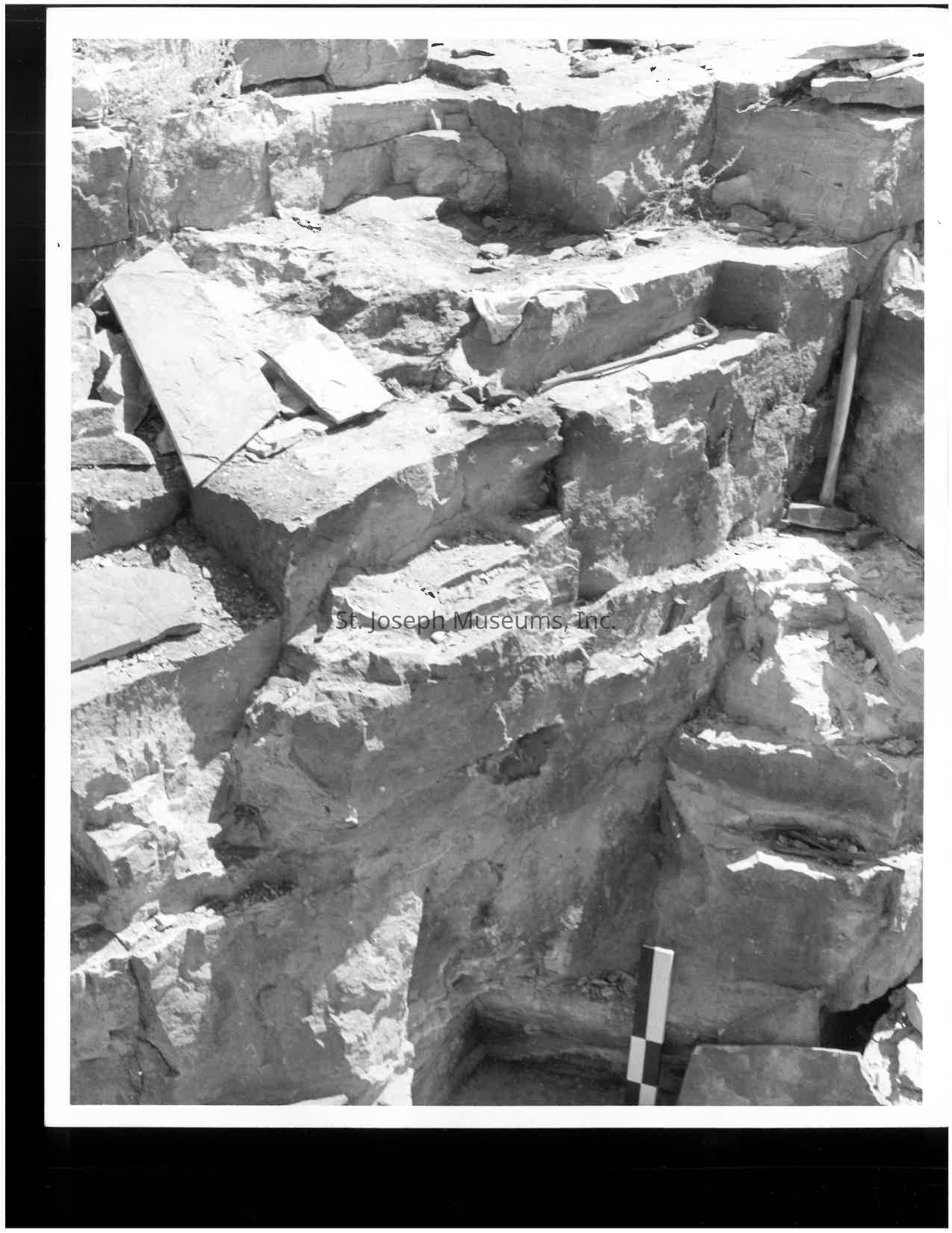 Click for PDF: The black and white image depicts a rocky excavation site featuring large, rugged stone formations with layered, jagged edges and a variety of tools scattered around. Sunlight casts stark shadows that accentuate the rough textures and contours of the rocks, highlighting the depth and complexity of the terrain. A measuring stick leans against one of the rock faces, indicating the methodical nature of the excavation process.
