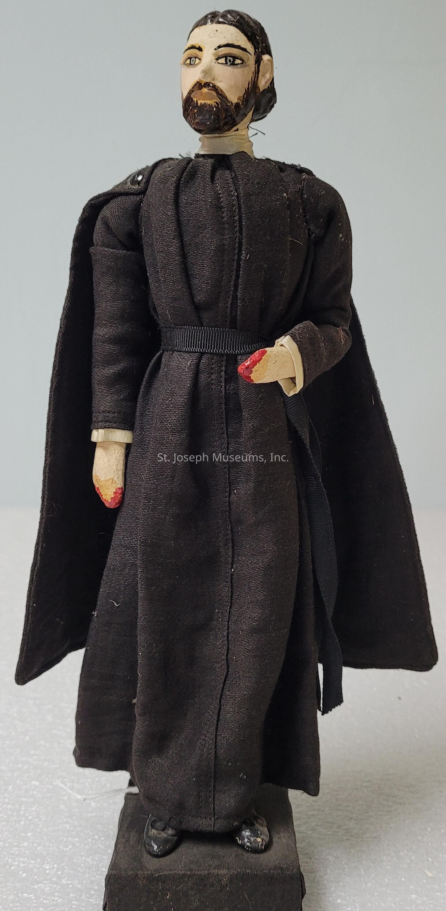 The image depicts a detailed handmade doll standing on a black rectangular pedestal. The doll is dressed in a long, dark robe with a matching cape and has intricately painted facial features, including a beard and expressive eyes. Its hands, which are shaped in a delicate manner, are also painted, with the right hand featuring red fingertips, possibly depicting nail polish or another form of decoration. The overall appearance of the doll is reminiscent of a historical or theatrical figure, lending it an air of dramatic presence. The textile of the robe has visible stitch lines, indicating meticulous craftsmanship. The combination of the dark clothing and the poised stance suggests a character of importance or mystique. Moreover, the doll's proportions and detailed painting contribute to a lifelike quality, despite its evident handcrafted nature. The pedestal base adds stability and enhances the display aspect of the doll, making it suitable as a collectible or decor item that captures attention through its intricate artistry and unique character representation.
