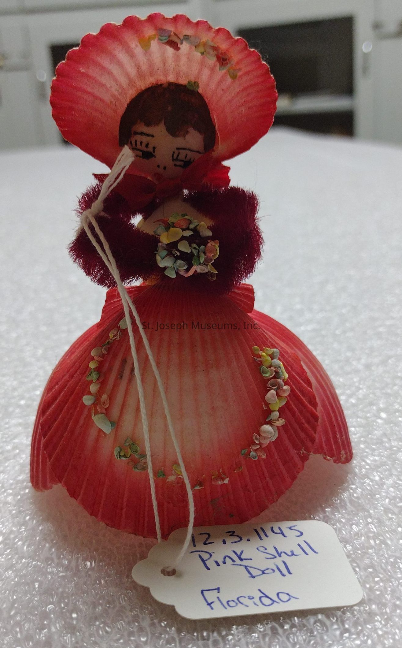 The image shows a decorative doll crafted from pink sea shells, with a scallop shell forming its main structure. The face of the doll is painted onto one of the shells, featuring closed eyes and simple facial details, with black hair drawn above. The doll is adorned with colorful, small embellishments and a red fluffy material forming a collar or part of its attire. It stands on a surface, likely made of soft material like bubble wrap. Attached to the doll by a string is a white tag with handwritten text, which reads "12.3.1145 Pink Shell Doll Florida," suggesting the item's name and origin.