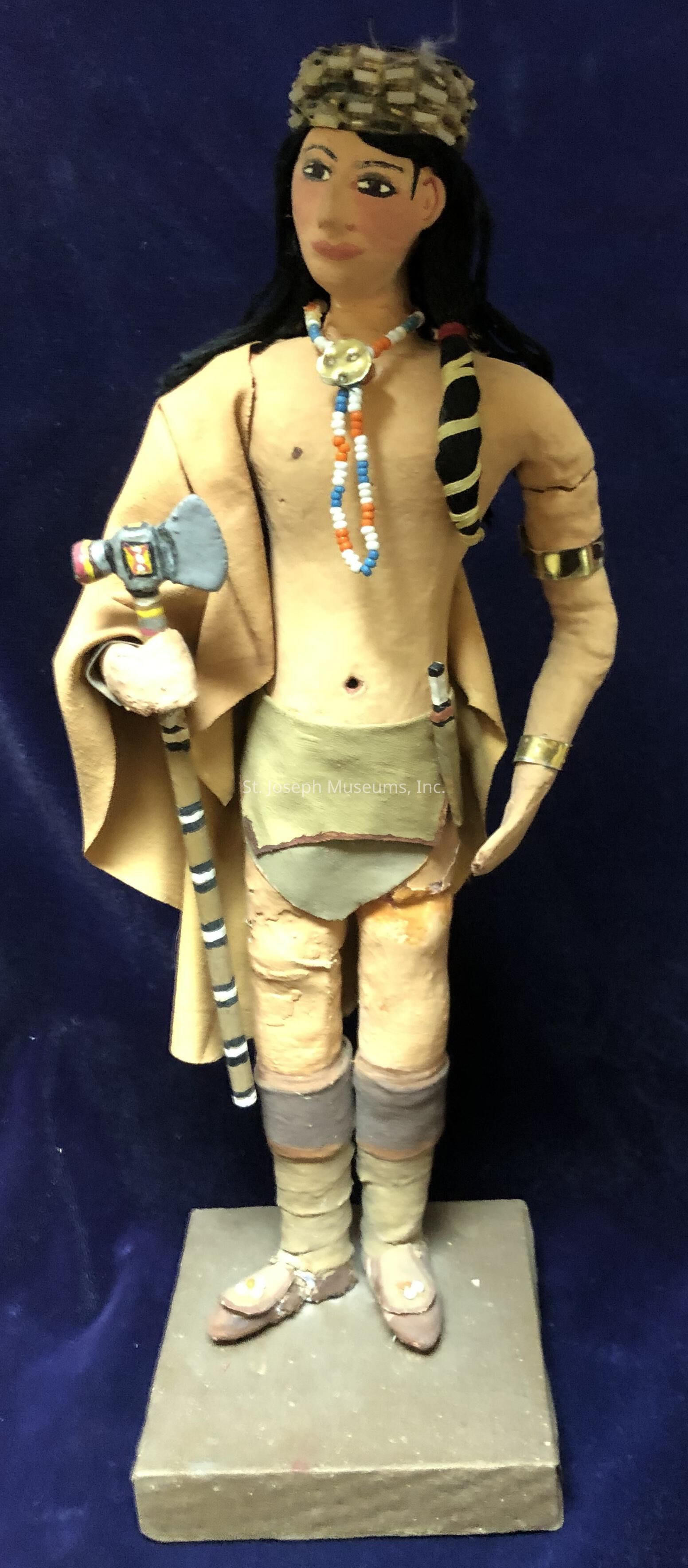 The image depicts a handcrafted figurine of a Native American man standing on a rectangular wooden base. The figurine is dressed in traditional attire, which includes a light brown vest and a short loincloth, complemented by knee-high leg coverings. His head is adorned with a woven headband, and around his neck hangs a multicolored beaded necklace. In his right hand, he holds an intricately detailed tomahawk, with a striped handle and colorful design on the blade. The figurine’s hair is depicted as long and dark, cascading over his shoulders. His attire and accessories capture the cultural essence of indigenous craftsmanship, and a beaded arm band decorates his left arm. The overall craftsmanship of the figurine reflects an artistic representation of Native American attire and tools, focusing on authentic elements like the beaded necklaces and the traditional weapon. The figure stands against a dark blue background, which contrasts with the earthy tones of the attire, emphasizing the figurine's detailed features and traditional motifs, lending