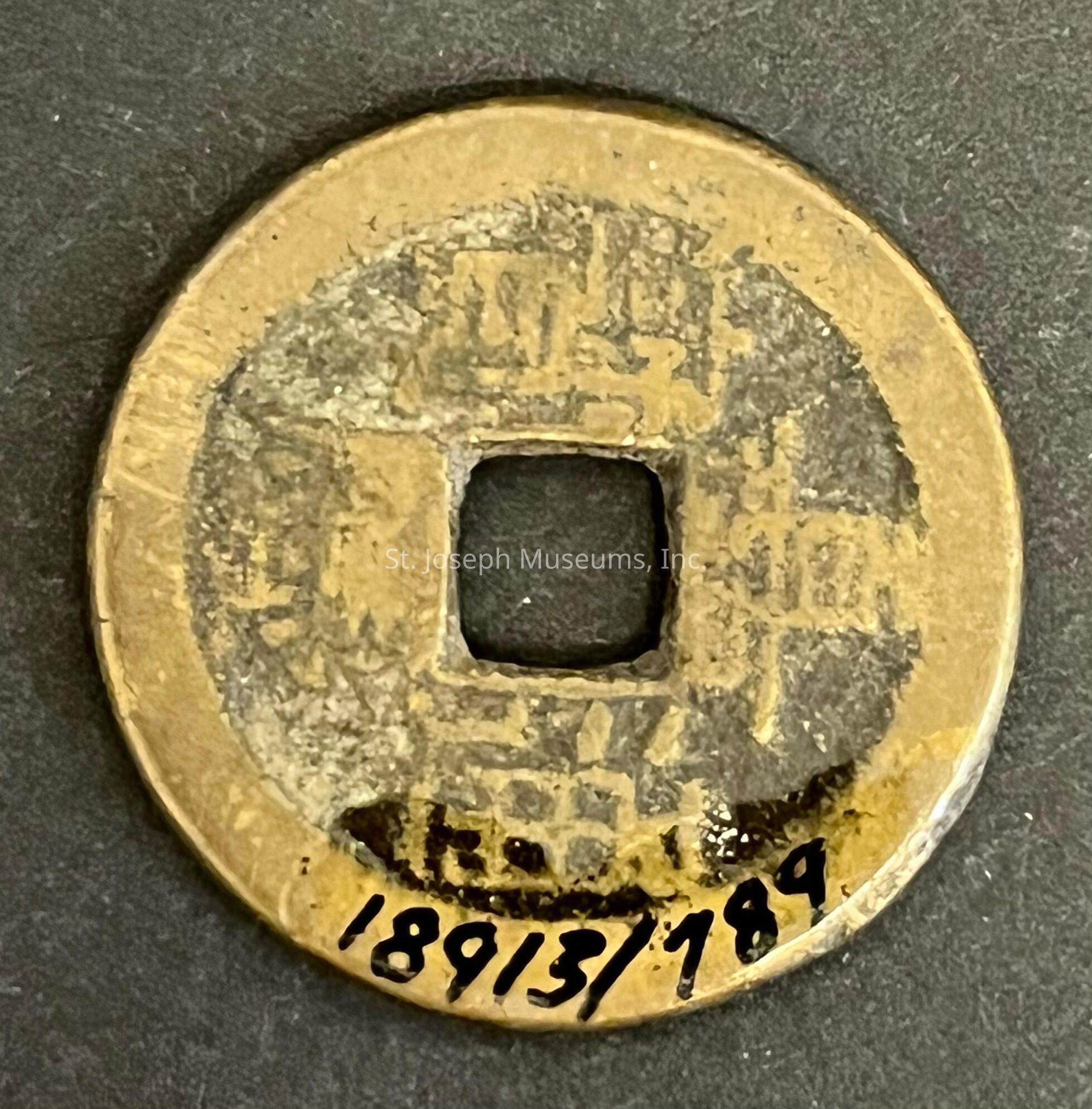 The image depicts an ancient round coin with a square hole in the center, featuring worn-out engravings of Chinese characters around the center on one side, indicative of its historical significance and cultural origin. The coin's metallic surface shows signs of aging and wear, characterized by a tarnished patina, which suggests it has endured a long passage of time and handling. Marked with a prominent set of numbers "1893/189" in black ink along one edge, the artifact is possibly cataloged for collection or exhibition purposes. The coin rests against a plain, dark background, further emphasizing its historical features.