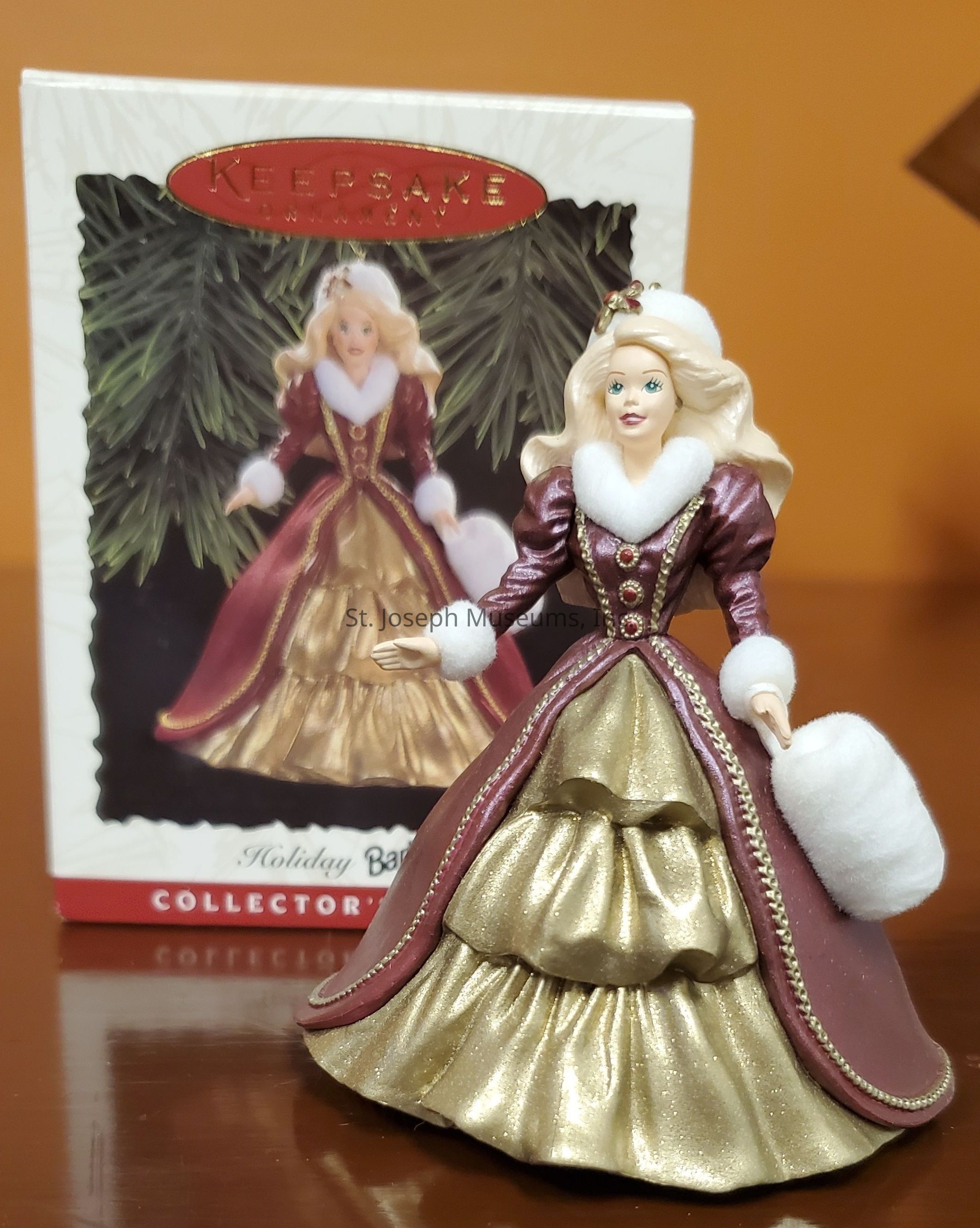The image features a detailed figurine of a woman dressed in an elegant, festive gown characterized by a luxurious deep burgundy color complemented by shimmering gold accents. The dress is adorned with white fur trim around the collar and sleeves, enhancing its wintry appearance. The figure holds a fluffy white muff in one hand, exuding a sense of vintage charm. Her long blonde hair cascades down gracefully, adding to her poised demeanor. In the background, a box with a similar image of the figurine can be seen, indicating that this is a collectible holiday keepsake, enhancing its appeal for decorative or collectible purposes.