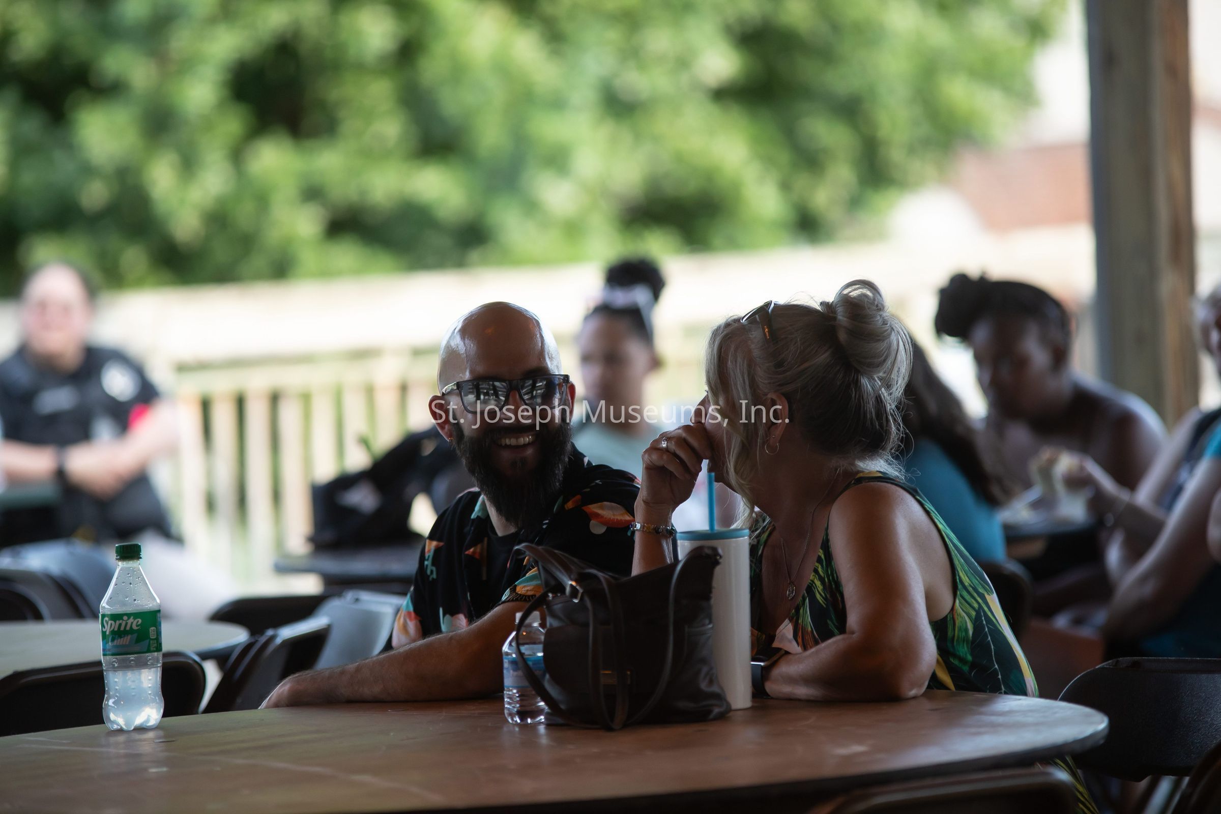 In this image, a man and a woman are sitting at an outdoor table, engaged in a lively conversation while smiling at each other. The man, wearing glasses and a shirt with a colorful pattern, looks directly at the woman with amusement. The woman, with her hair tied up and wearing a green-patterned dress, rests her chin on her hand with a playful expression. In front of them, there are various items, including a large cup with a straw and a bottle of Sprite placed on the table. The background features blurred figures of other people sitting at tables, along with trees and part of a wooden railing, suggesting a relaxed, casual atmosphere at an outdoor event or gathering.