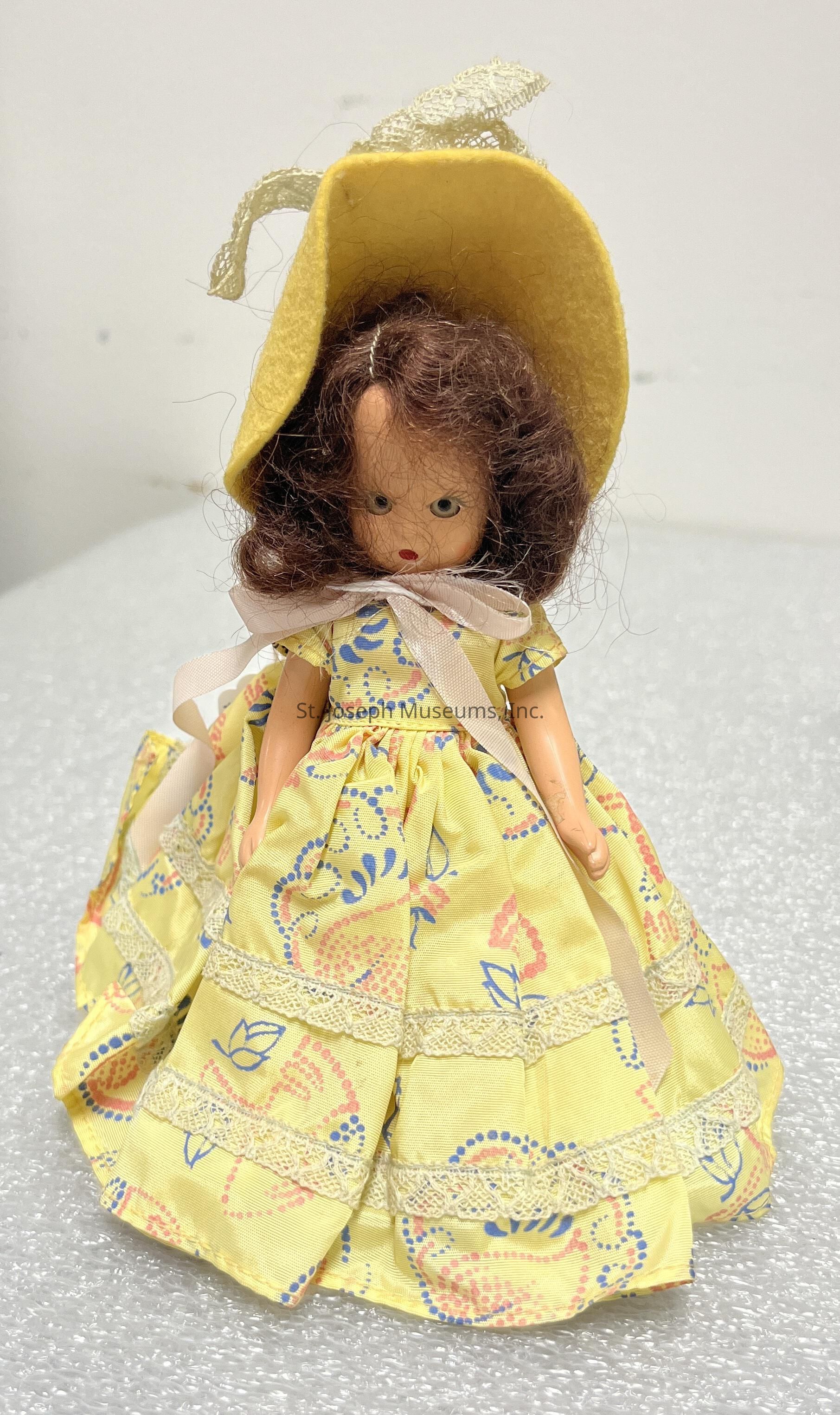 The image features a vintage doll adorned in a charmingly intricate outfit. She wears a yellow bonnet atop her curly brown hair, adding a quaint, old-fashioned appeal. Her dress, also predominantly yellow, is accentuated with pastel blue and pink floral patterns. The dress is decorated with delicate lace trims, providing an elegant and detailed appearance. A light-colored ribbon ties neatly at the neckline, further enhancing the doll’s classic look. The surface on which she stands appears to be a textured, light grey material, possibly resembling foam or fabric, providing a neutral backdrop that highlights the doll’s vibrant colors and intricate details. Her facial expression is neutral, with painted features that include red lips and deep, contemplative eyes. This doll exudes a sense of nostalgia and timeless charm, reminiscent of handcrafted toys from a bygone era. Each element of the doll's attire suggests careful craftsmanship, evoking imagery of dolls cherished and lovingly played with in historical domestic settings.
