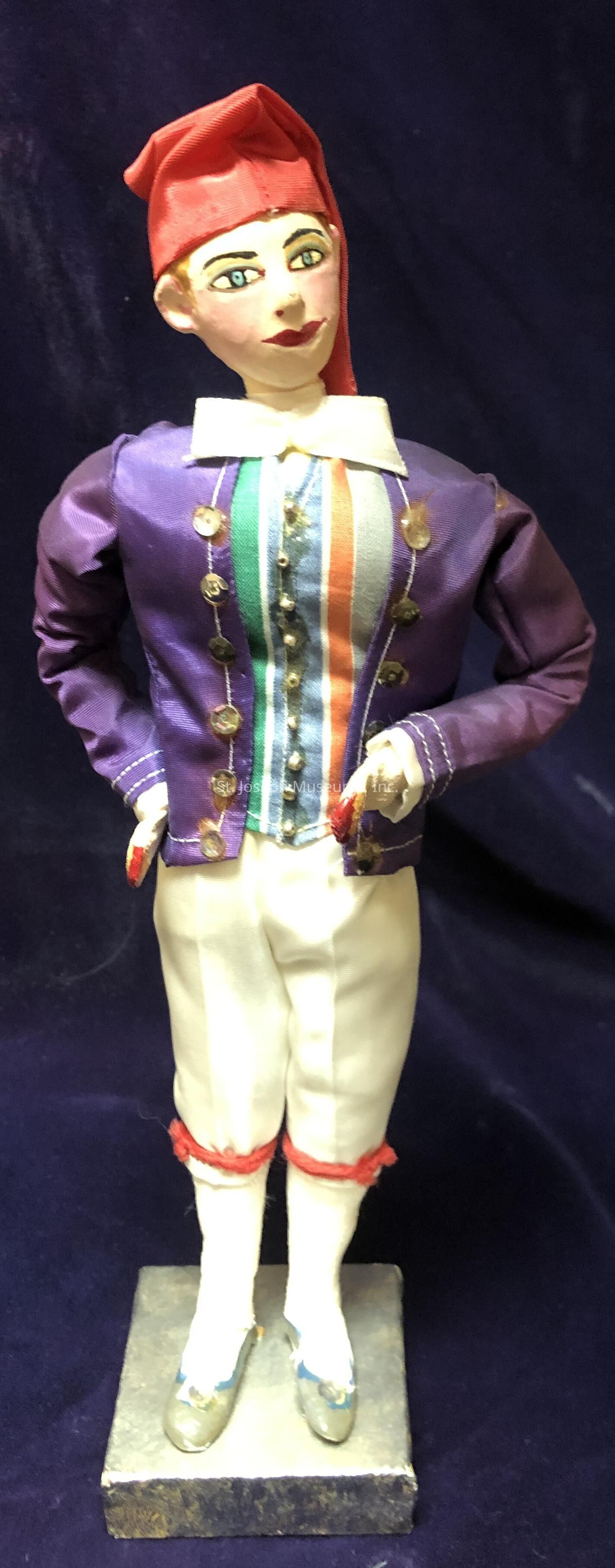 The image features a detailed figurine dressed in a colorful, theatrical costume, reflecting a whimsical and playful style. The figure wears a vibrant, deep purple jacket adorned with gold buttons, giving it a festive appearance. Underneath the jacket, the figure has a shirt with vertical stripes in various colors, adding to the lively and dynamic look. A white collar peeks out from the top of the jacket, contrasting with the deeper tones. The pants are white, with red trim around the knees, giving a traditional yet cheerful touch. On their feet, the figure dons stylish shoes with small decorations, resting on a sturdy base. The person’s head is embellished with a bold, red hat that complements the ensemble. The background is a rich, dark fabric, which highlights the figure’s vibrant attire, creating a visually striking and engaging composition that evokes a sense of performance or festivity, possibly reminiscent of a circus or theatrical setting.