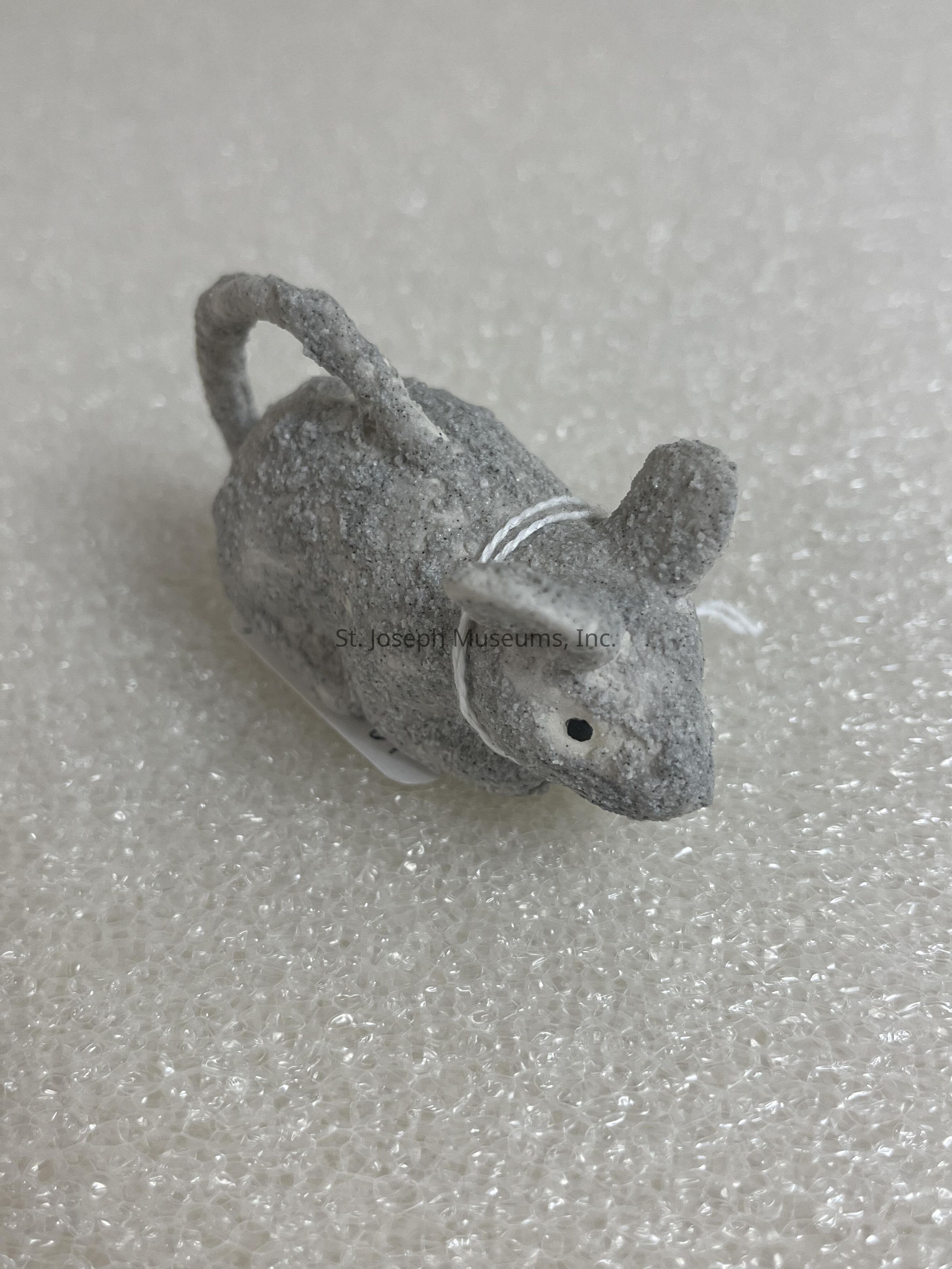 The image displays a small, textured figurine of a mouse standing on a surface that appears to be bubble wrap or a similar textured material. The mouse is gray and has a slightly rough texture, resembling a handmade or artisanal craft object. Its tail is curled upward, and there's a thin white string looped around it, suggesting it might be designed as an ornament or decorative item. The surface beneath provides a contrasting background that highlights the details of the mouse figurine, including its ears, nose, and simple black eye dot. The lighting in the image is bright, emphasizing the texture of both the figurine and the surface.