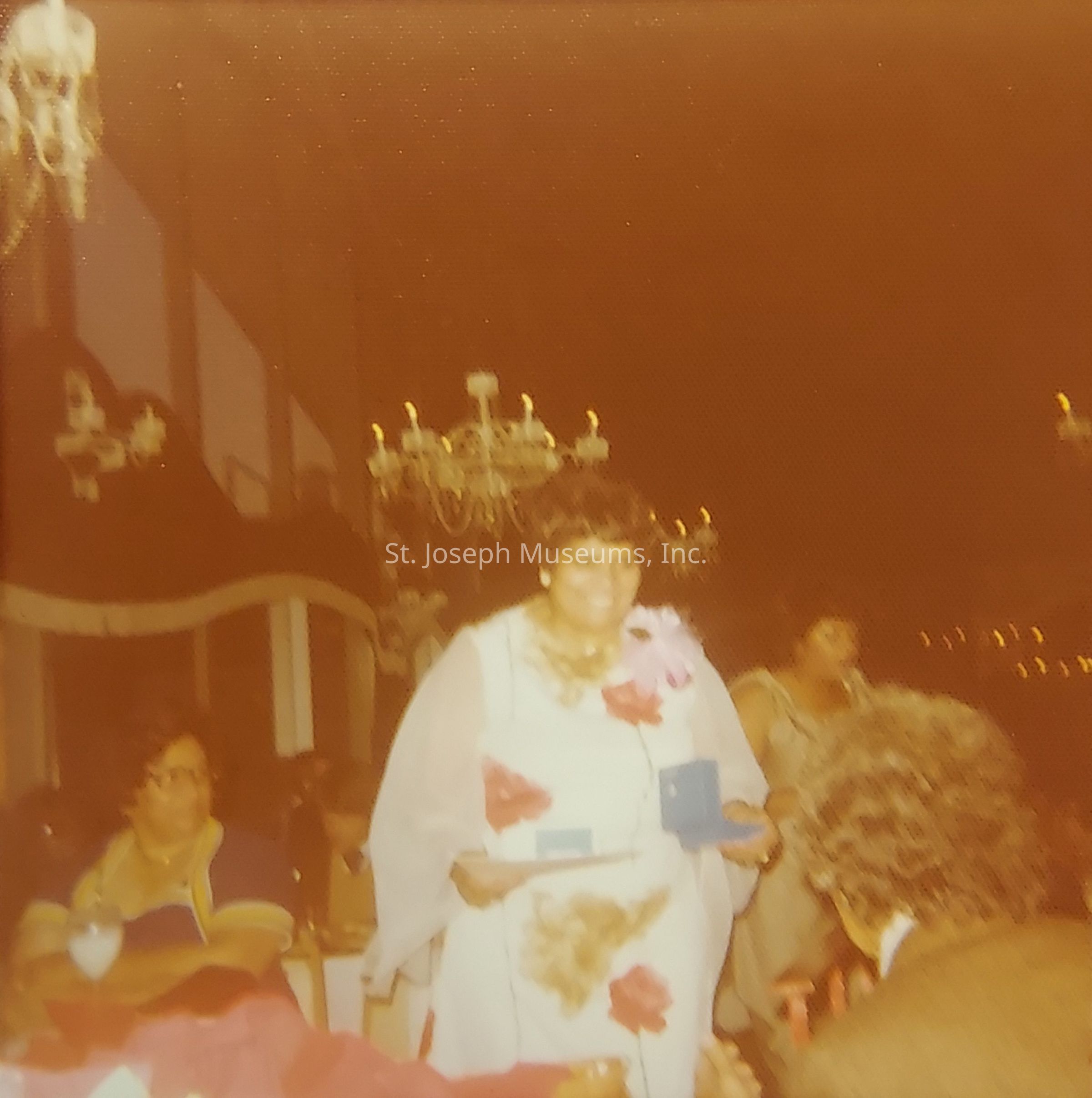 The image captures a warmly lit indoor scene where a woman wearing a white dress adorned with red and brown floral patterns, complemented by a sheer shawl and a large pink flower on her shoulder, is standing among a gathering of people at what appears to be a formal event; she is holding a small blue box and a piece of paper with a smile, drawing attention amidst the elegant surroundings featuring ornate chandeliers and numerous attendees engaged in conversation, two of whom are visible in the background, one seated and observing the proceedings while another stands nearby, adding to the sophisticated ambiance of the occasion.