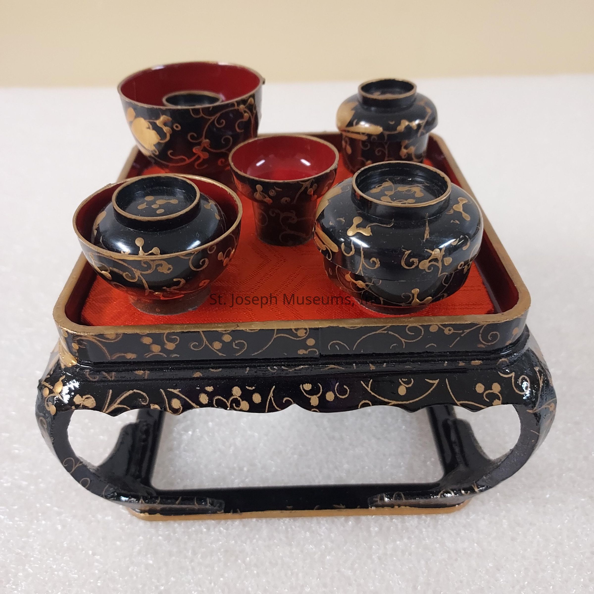 The image depicts an elaborate lacquerware set consisting of five intricately designed bowls and vessels, each adorned with golden patterns on a black background, placed on a small lacquered tray with elegant curved legs. The interior of the vessels and the surface of the tray are painted a rich, deep red, creating a striking contrast with the black and gold exterior, with swirling motifs enhancing the overall aesthetic. The craftsmanship showcases traditional artistry, reflecting a sense of elegance and cultural heritage, likely used in ceremonial or special occasions to highlight its significance and the skill involved in its creation.