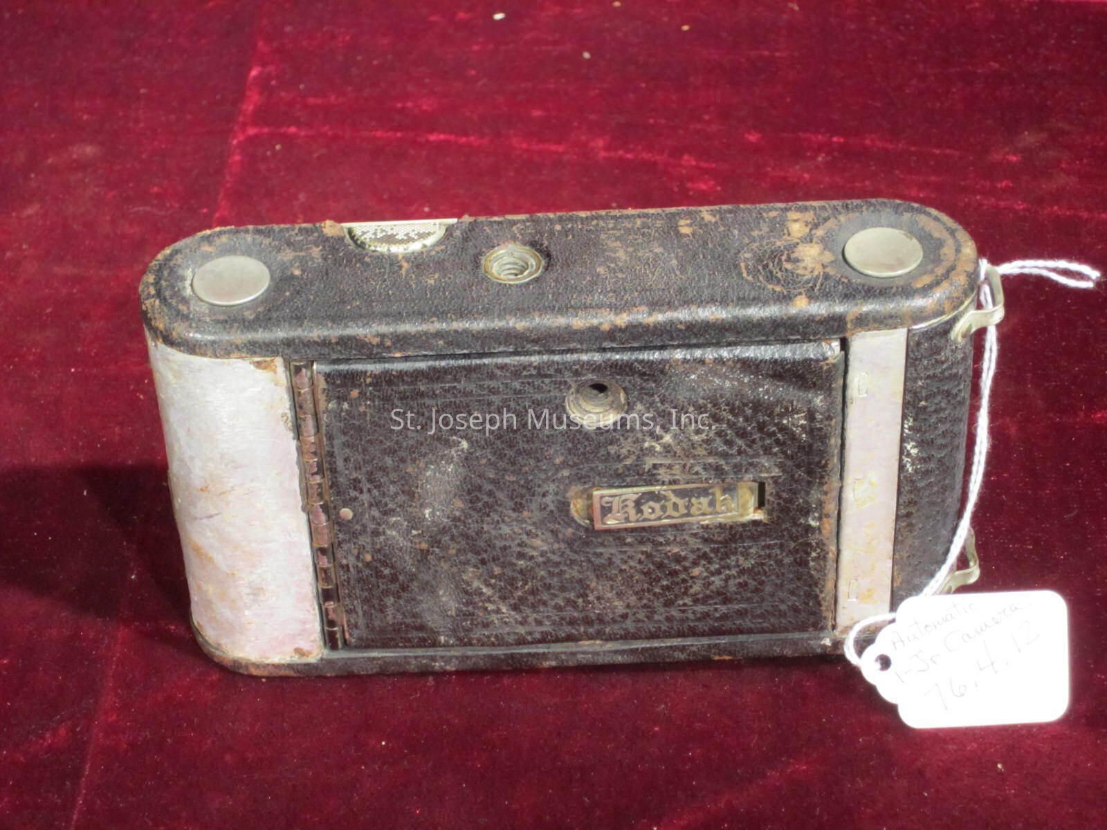 The image displays a vintage Kodak folding camera placed on a rich, textured red surface. The camera has a worn appearance with a black leatherette covering, showing signs of age and use, with metal accents visible at the ends. The front panel bears the Kodak emblem, indicating its brand, while a white tag attached with string provides handwritten information, possibly for cataloging or sale purposes. The camera, with its compact and rectangular shape, suggests it was designed for portability and convenience, reflecting the historical charm and mechanical ingenuity of early photography equipment.