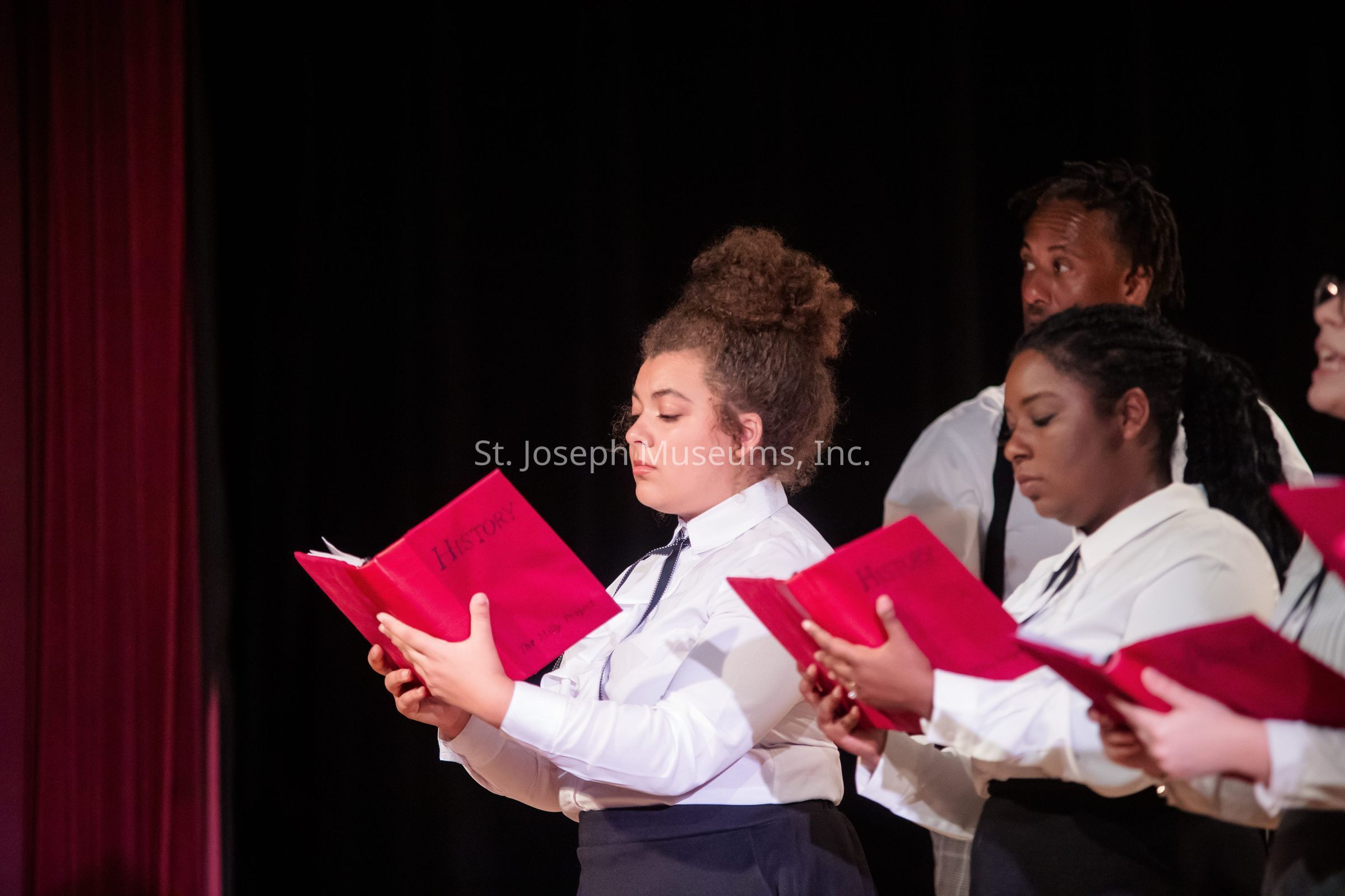The image depicts a group of individuals dressed in formal attire, holding and focusing on bright red books titled "History," participating in what appears to be a choir or singing event, likely on a stage with a dark background and red curtains, suggesting a solemn or formal recital or performance.