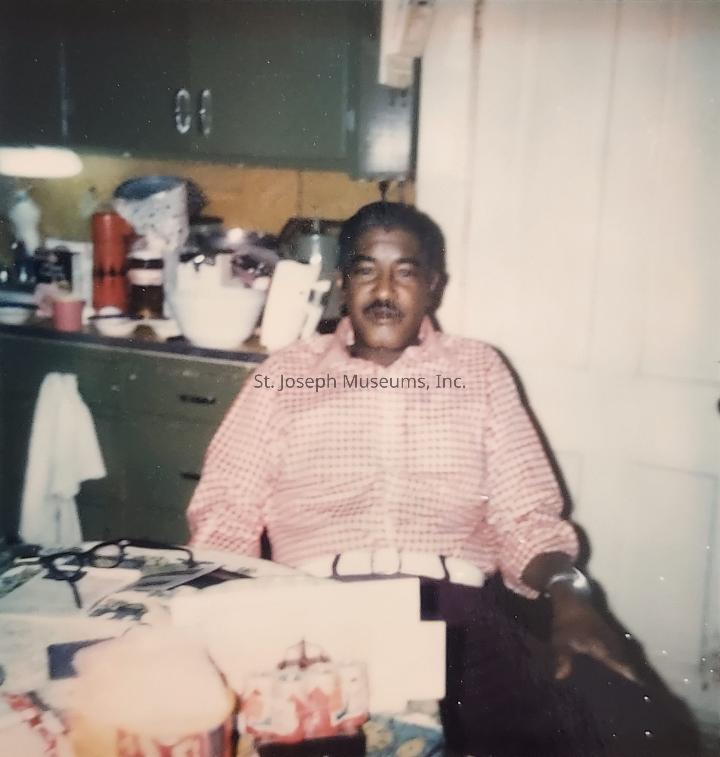 The image features a man seated at a kitchen table, wearing a pink and white checkered shirt. Behind him, the kitchen has green cabinets filled with various household items like bottles, bowls, and a towel hanging on a drawer. On the table in front of him, there are glasses, boxes, and cans, possibly of soda. The man's expression is calm and relaxed as he situates himself in what appears to be a warm and lived-in atmosphere, evoking a sense of everyday normalcy and comfort. The lighting and color tones suggest it might be an older photograph, adding a nostalgic feel to the scene.