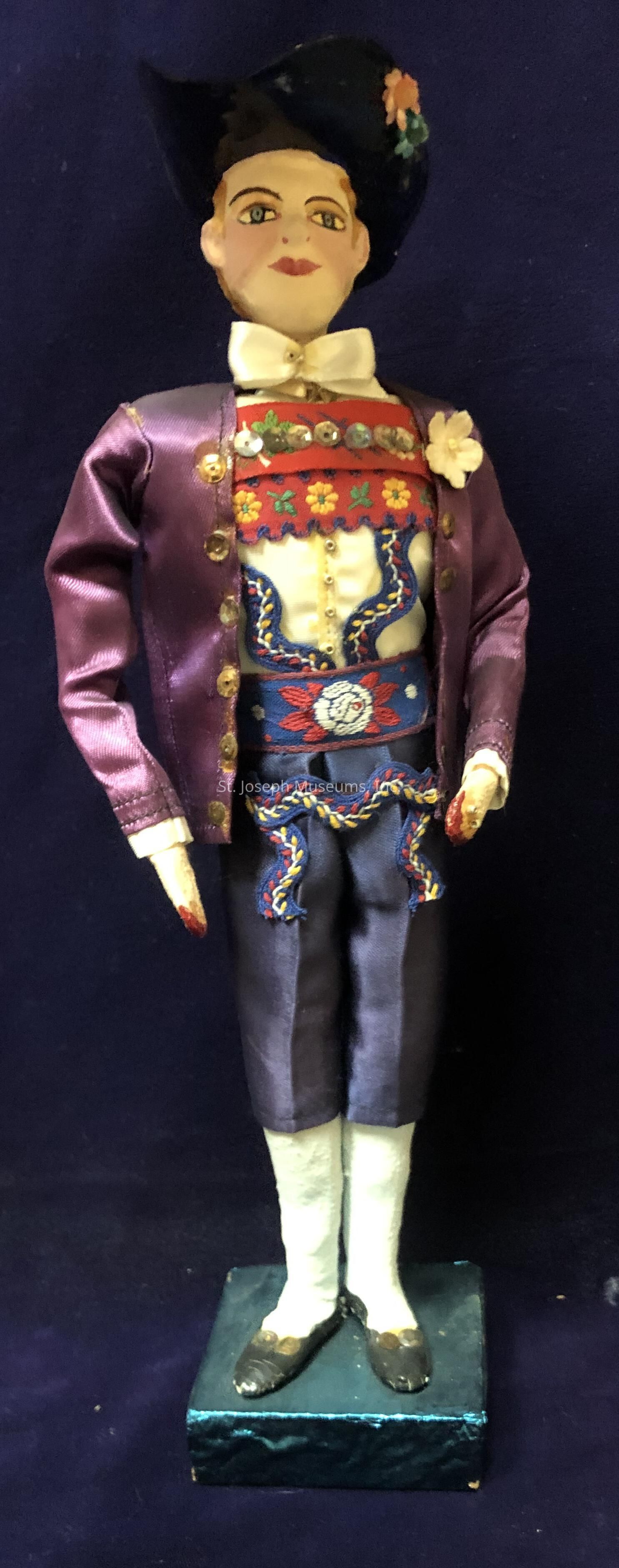 The image features a intricately detailed doll dressed in a traditional attire, standing on a small pedestal. The doll wears a purple satin jacket adorned with gold buttons, over a vividly embroidered vest showcasing floral patterns in vibrant colors such as red, yellow, and blue. The white collar and bow tie provide a refined contrast to the colorful motifs. The dark hat with floral embellishments atop the doll's head adds to its elegant appearance. The trousers, a deep shade of blue, feature additional embroidery along the sides, complementing the overall design. The doll’s shoes, with a subtle shine, rest on a textured base, and the entire setup is against a deep navy background, emphasizing the bright colors of the attire. This decorative doll appears to be a representation of cultural or historical significance, meticulously crafted to capture the essence of traditional garb.
