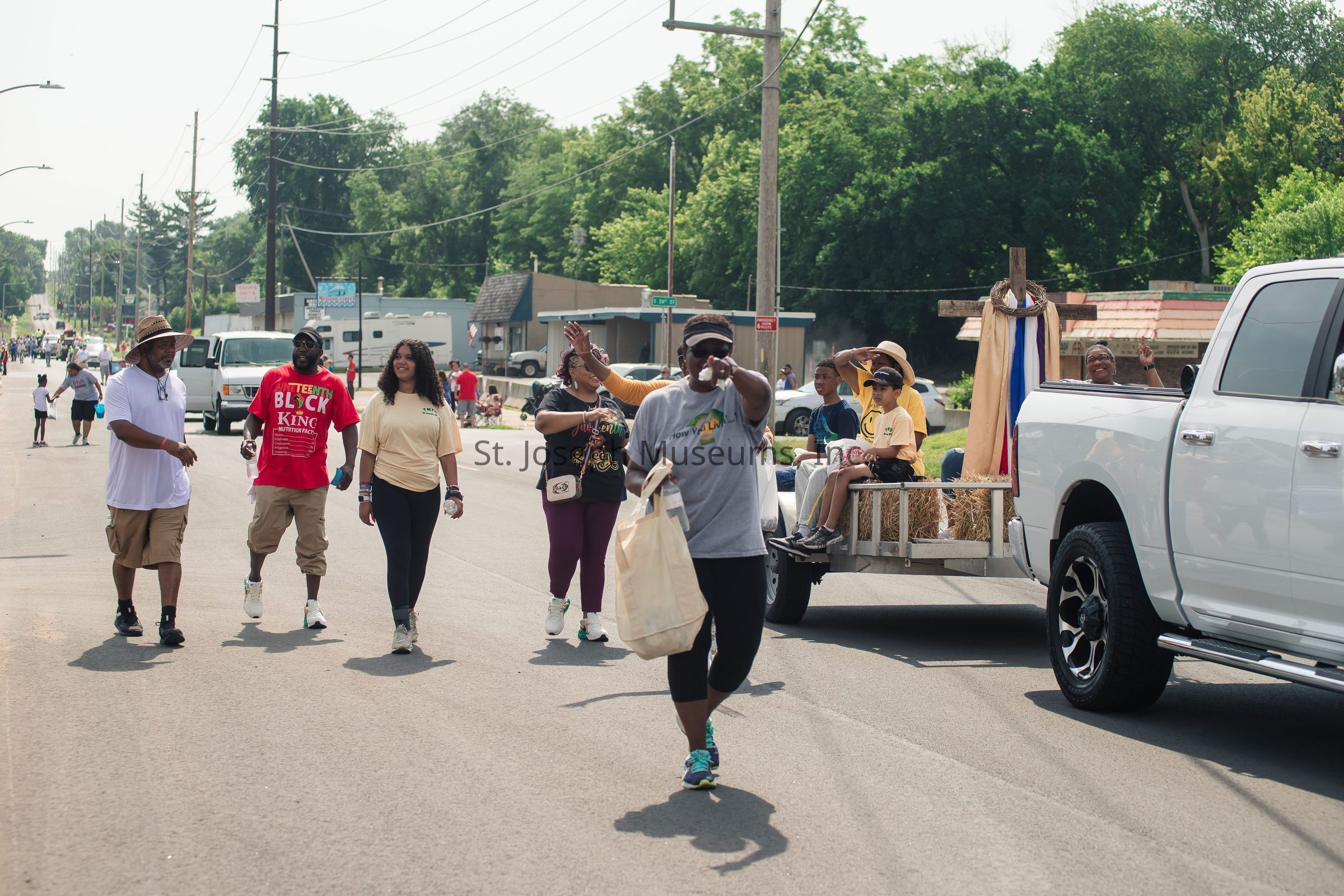 The photograph captures a lively street scene where a group of people is participating in a parade. In the foreground, a person wearing a gray shirt is pointing towards the camera while carrying a tote bag, leading others who walk behind them. Participants, dressed in casual attire, show a joyful demeanor, with one individual wearing a t-shirt that reads “TEAM BLOCK KING.” On the right, children and adults are seated on a float constructed from hay bales, pulled by a white truck, with some waving cheerfully. The background displays a typical suburban street with trees, utility poles, and various small buildings, suggesting a community celebration on a warm day.