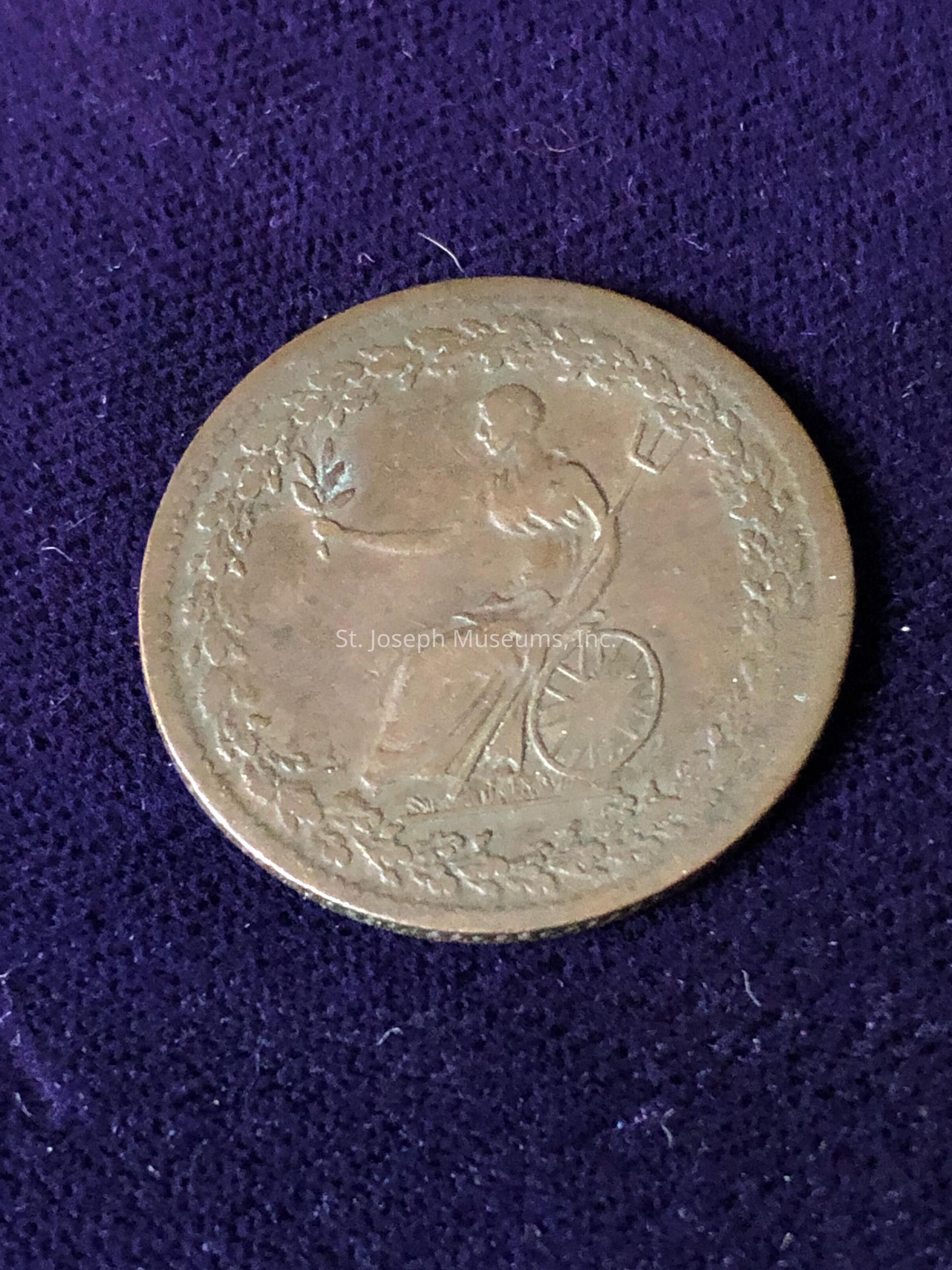 The coin features an intricate design with a central figure seated on a chariot, possibly representing a classical deity or symbolic figure. The figure is holding an olive branch in one hand, and there seems to be an agricultural tool, like a trident, in the other hand. Surrounding the figure is a detailed, decorative border of leaves or laurels, adding a sense of elegance and historical significance to the coin. The coin's surface appears worn, suggesting it has aged over time.