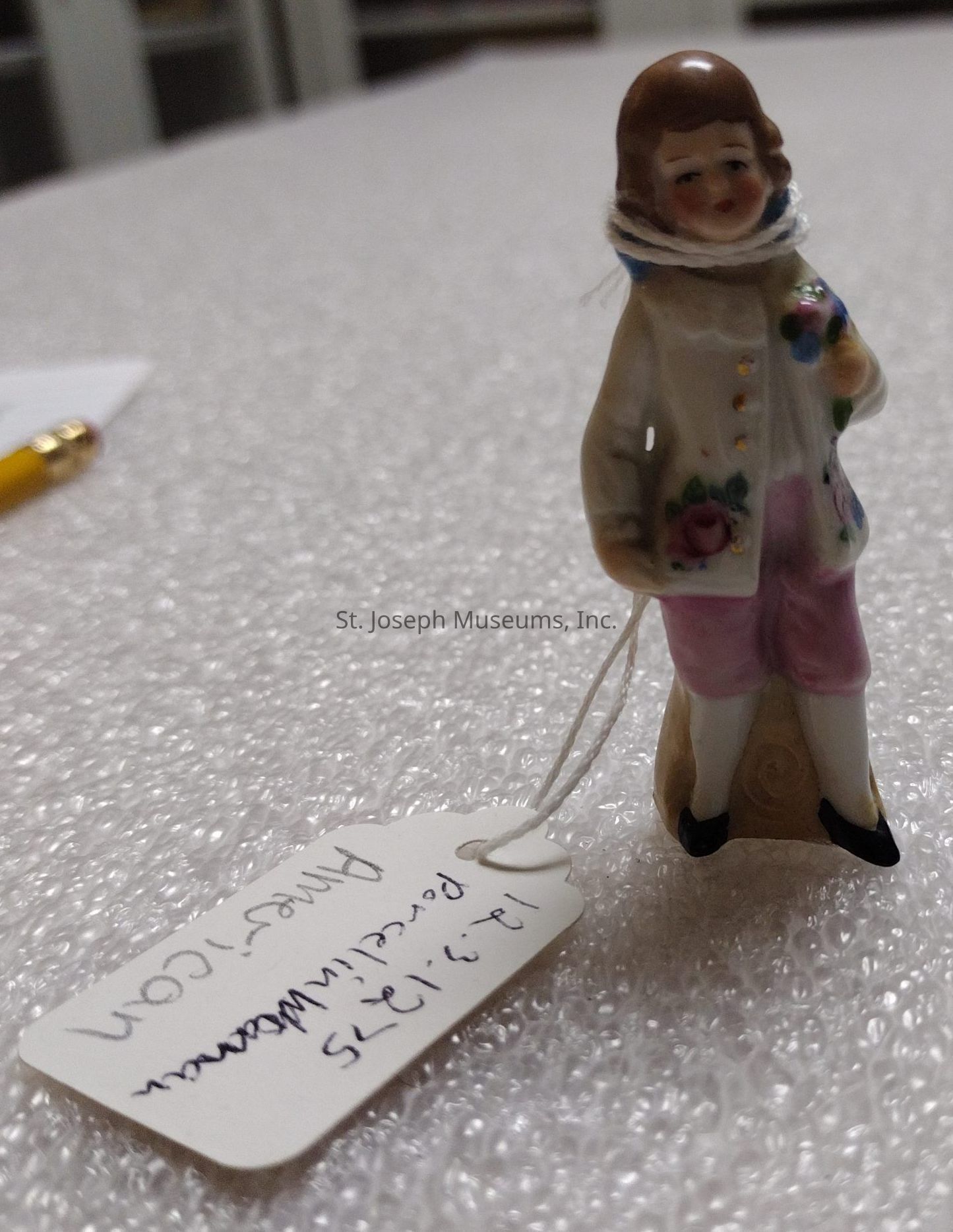 The image depicts a small porcelain figurine of a person wearing a cream-colored jacket adorned with floral designs, pink trousers, and black shoes, holding a bouquet of flowers. The delicate figurine is tied with string to a white tag that appears to have some handwritten notes on it, indicating "Porcelain Figurine" along with a date, "13-9-1375". The figurine is set against a textured surface that resembles bubble wrap, suggesting it may be in a setting related to storage or transportation. In the background, there is a hint of a pencil, further implying the setting might be part of an inventory examination or packing process. The intricate details, such as the facial expression and the careful painting of the flowers, indicate craftsmanship.
