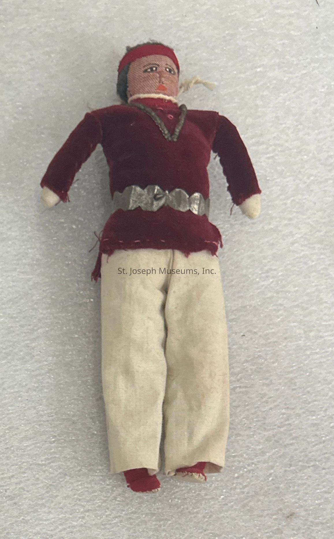 The image shows a handcrafted doll lying on a textured white surface, wearing a deep red top with long sleeves and a matching headband adorned with a metallic, decorative strip around the waist. The doll's cream-colored pants extend to its red-footed base, with minimalist facial features that are stitched on, including small eyes, a nose, and lips. Additionally, the doll appears to have short, dark hair made from fibers, and its overall appearance evokes a sense of traditional craftsmanship, likely reflecting cultural attire or historical styles, adding character and charm to this unique piece. The doll's left arm, slightly bent, contrasts with the right arm, which seems more elongated, both constructed from a fabric that complements the color palette. This balance of hues, materials, and intricate detailing suggests a focus on handmade artistry that tells a story, inviting observers to ponder its origins and the hands that lovingly crafted this soft, expressive figure.