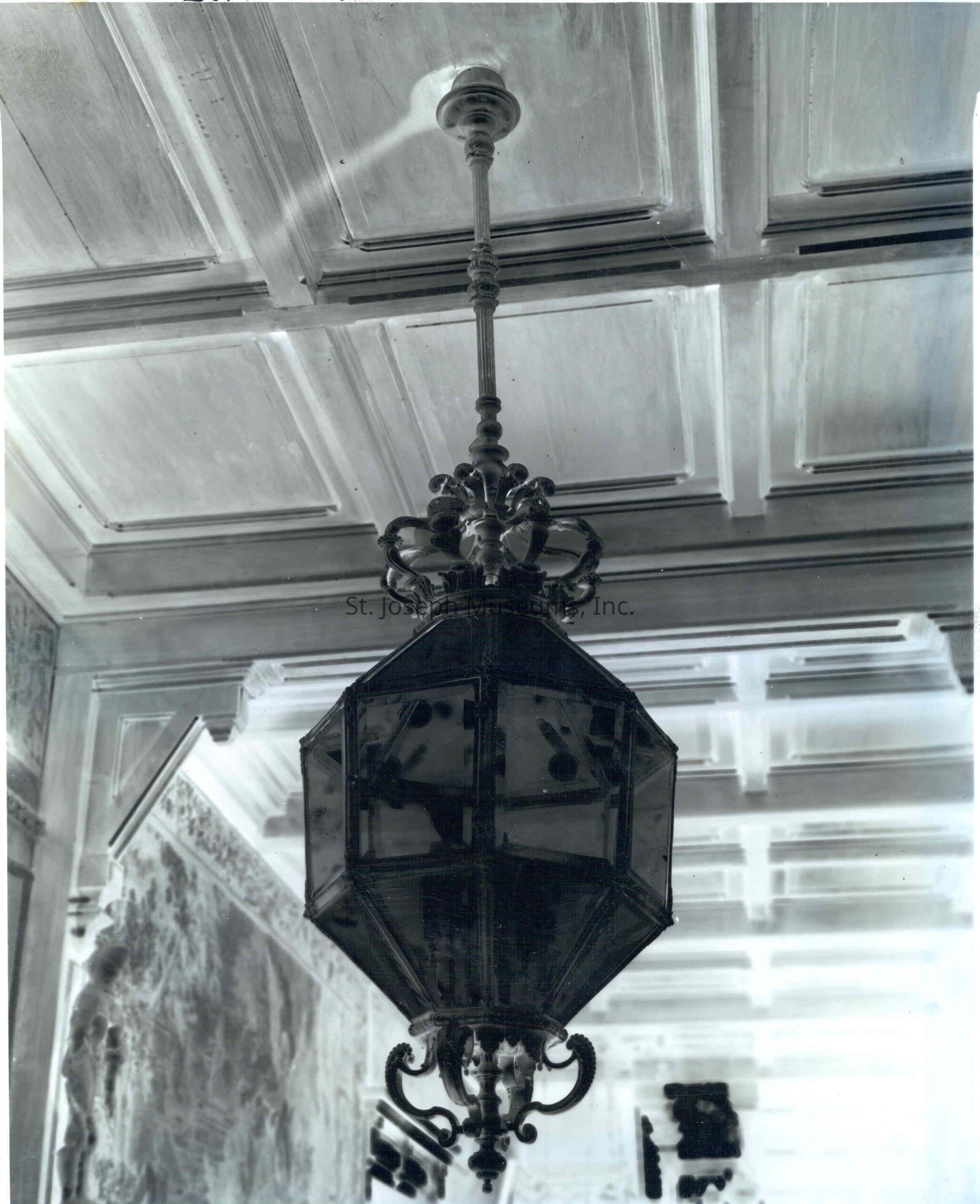 Tootle Mansion Front Hall Chandelier, c. 1900: Copyright: 84.21.1902 Tootle Mansion Front Hall Chandelier, c. 1900
The negative depicts a close-up of a chandelier hanging in the front hall of Tootle Mansion, dating back to around 1900. The chandelier is detailed and ornate, with an intricate metal design. Above, the ceiling is tiled in a geometric pattern with coffered panels.