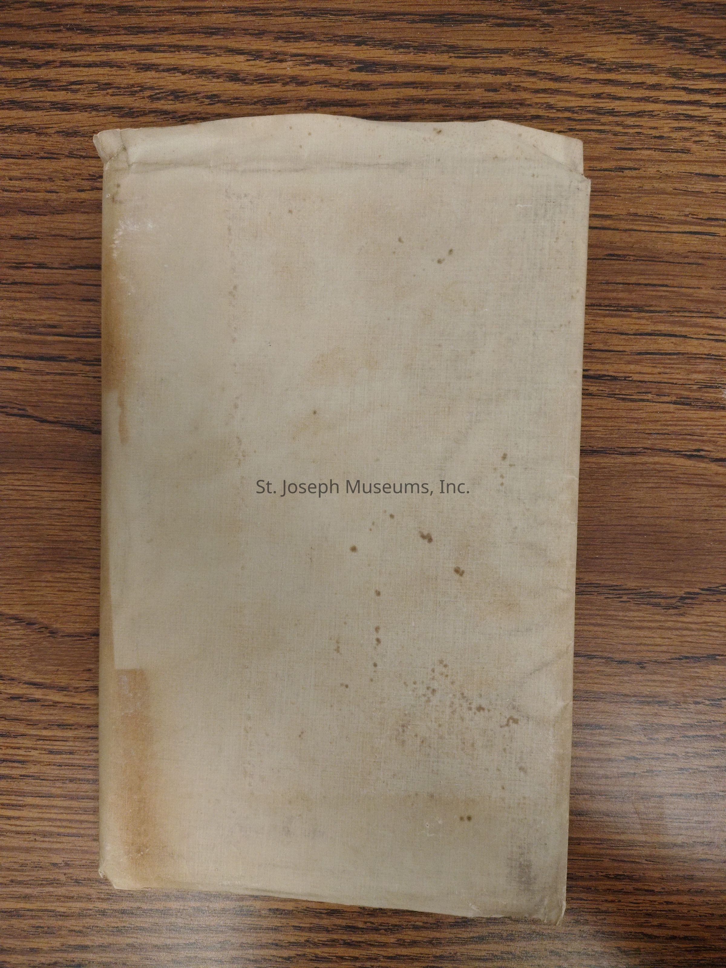 The image depicts a closed, aged book laying on what appears to be a wooden surface with a rich, brown texture. The book's cover, made of a cloth or paper material, exhibits noticeable signs of aging, such as discoloration, fading, and various dark stains and spots scattered across the surface. The cover is a light, beige color, suggesting it may have once been lighter before exposure to elements or use over time. The edges are slightly frayed, indicating significant wear and tear likely due to handling and passage of time. The texture of the cover also shows a somewhat coarse and papery appearance, which could suggest it’s a fabric-covered book rather than a leather one. The book’s spine appears slightly bent or curved, hinting at frequent usage or pressure from being stacked or stored with other volumes over many years. Positioned diagonally in the frame, the book’s placement is parallel to the vertical grain of the table's wood, creating a pleasing visual balance between the book and its resting