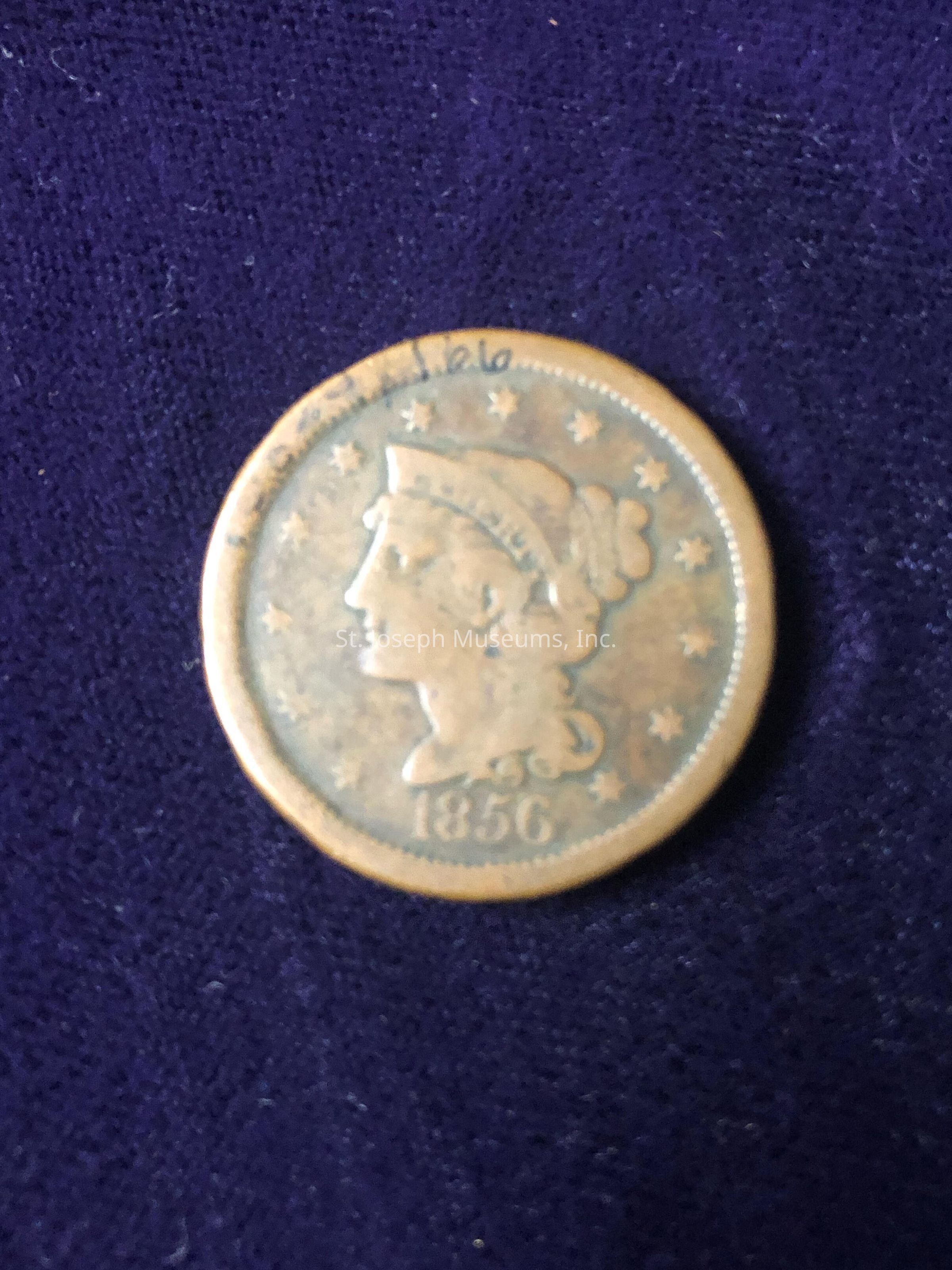 The coin appears to be a vintage piece from the year 1856, indicated by the date prominently displayed at the bottom. The obverse side of the coin shows a profile of a woman, possibly Lady Liberty, facing left. Surrounding the profile are thirteen stars, which is a common design element on many historical American coins, symbolizing the original thirteen colonies. The coin has a worn appearance, with discoloration and signs of aging, suggesting it might have been circulated extensively or poorly preserved. Despite its age, the intricate details, such as the woman’s hair and headdress, are still discernible. Additionally, the rim of the coin seems to have slight writing or marks that might have been added later, and its overall patina reflects a rich history, evoking curiosity about the coin's origin, use, and journey over the years. The deep purple cloth beneath provides a contrasting backdrop, highlighting the coin's antiquity