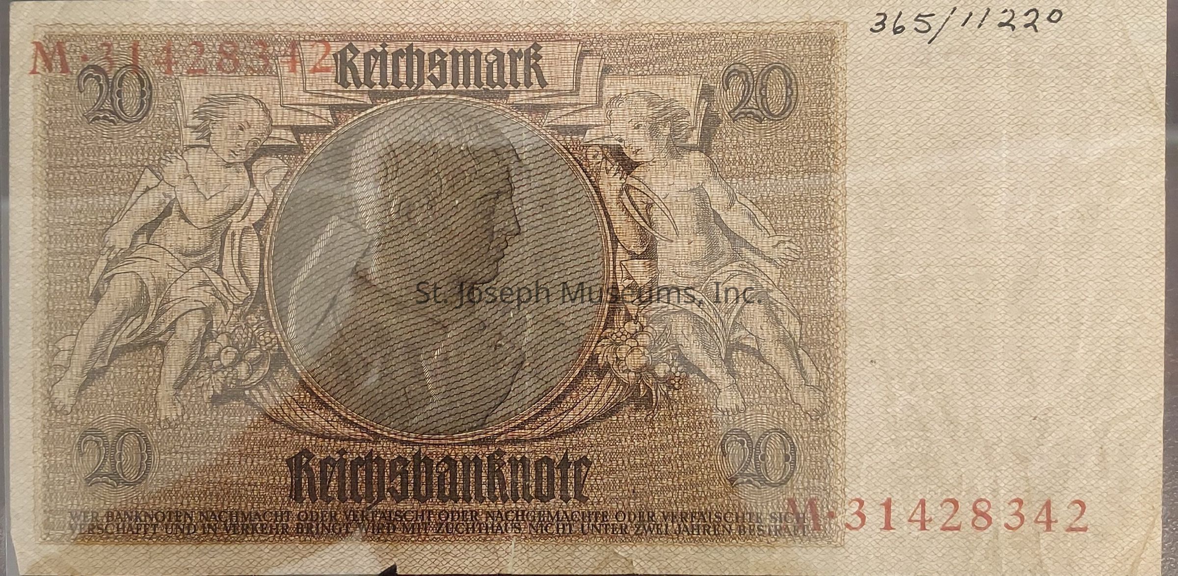 The image displays a vintage 20 Reichsmark banknote featuring intricate artwork and designs typical of old currency. Centered on the note is a prominent circular portrait, bordered by two cherubs or allegorical figures, each depicted with classical styling and seated atop decorative elements such as fruits and foliage. The denomination "20" is visibly marked in both top corners, and the word "Reichsbanknote" is emblazoned in an ornate, Gothic typeface across the bottom, alongside a serial number printed in red in the top left corner and bottom right corner.
