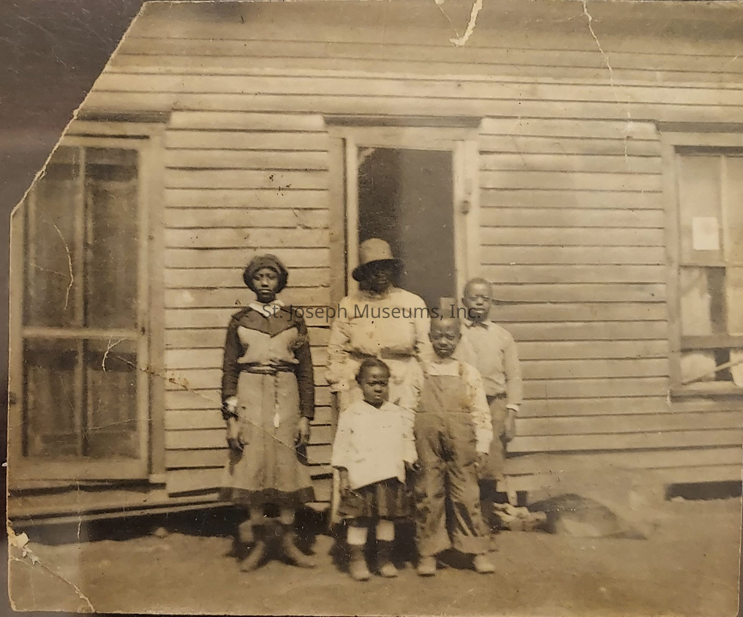 The image shows a vintage photograph of five individuals standing in front of a wooden house. The group includes an adult and four children, with varying expressions on their faces. The adult, wearing a hat, stands behind the children who are positioned in front, creating a balanced composition. The children are dressed in period clothing typical of the early to mid-20th century, featuring layers and practical materials. The house serves as a backdrop with visible details like a door and windows, adding context to the setting. The photo exhibits signs of aging, such as creases and a sepia tone, indicative of its historic significance. The mood appears solemn yet dignified.