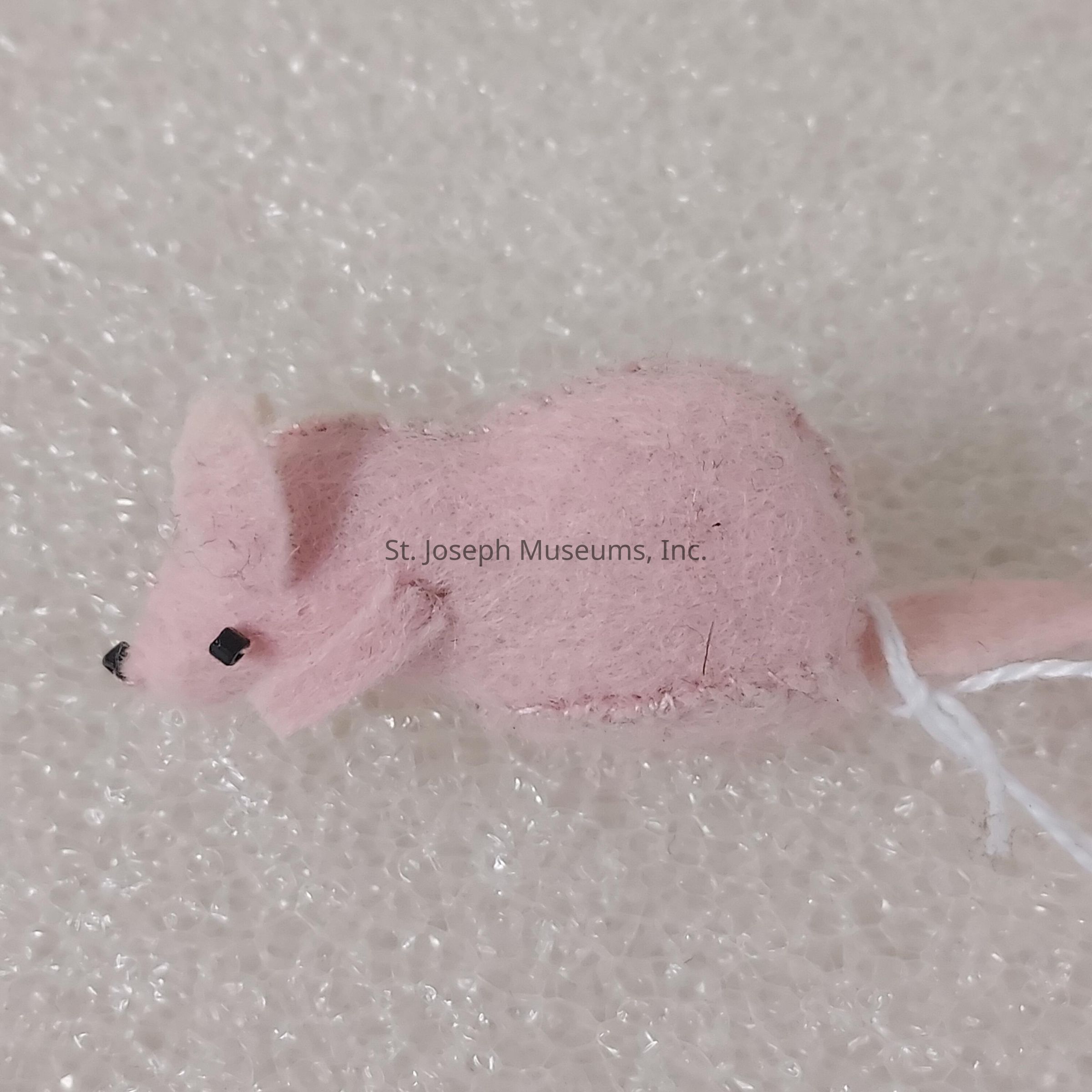 The image depicts a small, plush toy mouse crafted from pink felt material, featuring distinct details such as black eyes and nose, as well as large, rounded ears. The mouse has a lightly textured body, and its tail is made from the same pink felt but extends through a visible white string, suggesting a handmade or craft project feel. The background consists of a soft, white, foam-like surface that contrasts with the pink color of the toy, adding a tactile dimension to the image. The overall composition highlights the whimsical and cute nature of the felt mouse, making it appealing and charming.