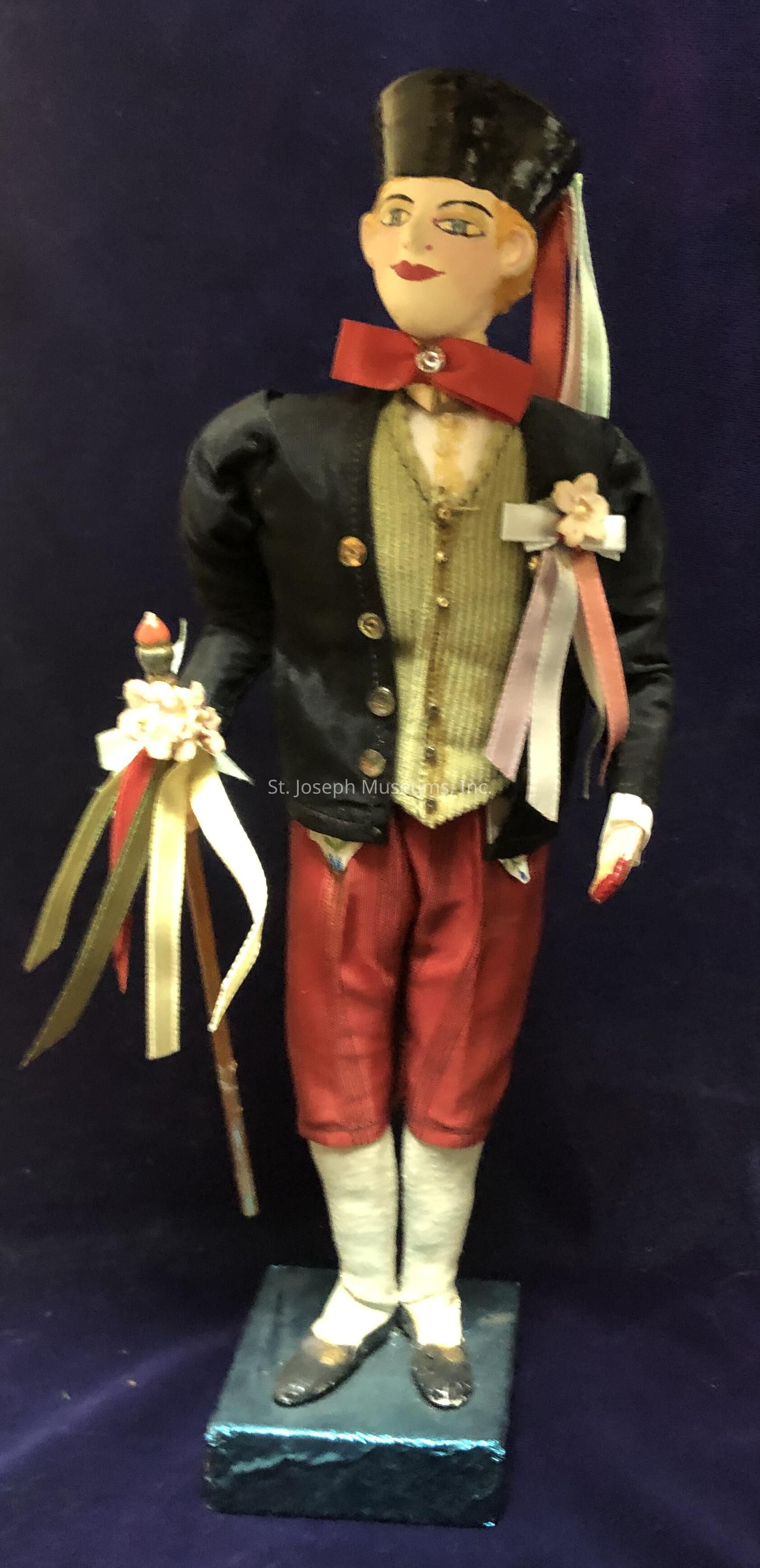 The image shows a handmade doll with a whimsical appearance, dressed in period-style clothing. The doll is wearing a black jacket with gold buttons over a textured vest, paired with red trousers. Its head is adorned with a tall black hat, and a striking red bow tie with a jewel detail completes the ensemble. The doll holds a stick adorned with colorful ribbons in one hand, and a floral decoration is attached to its chest, enhancing its festive look. It stands on a small pedestal against a dark background, giving it a charming and historic appeal.