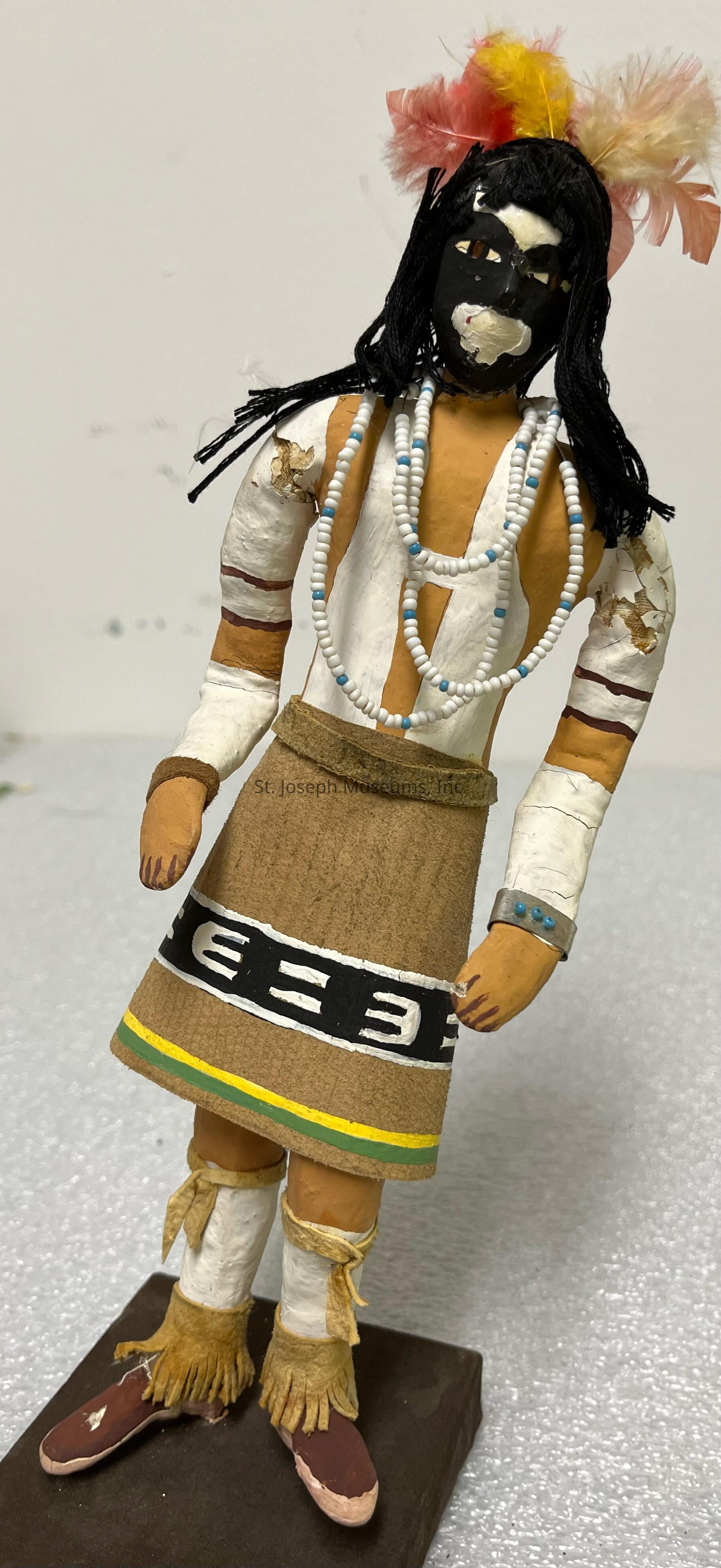 The image shows a handcrafted figurine, possibly a kachina doll, with a detailed design and culturally significant attire. This figure is characterized by a predominantly black-painted face with white markings and expressive features. Its head is adorned with vibrant feathers in shades of yellow, pink, and white, which add a colorful flair to the overall design. The figurine's long black hair contrasts with the bright beaded necklaces that drape over its shoulders and chest. It wears a short, rust-colored skirt with intricate geometric patterns, outlined by black and white belt-like detailing near the waist. The lower part of the skirt features yellow and green trim, providing more color accents. The arms and legs have painted designs, including white bands intersected by brown, matching the arm decorations. Leather fringes and traditional moccasin-style sandals complete the attire. The entire figure stands on a flat brown base, suggesting it is intended for display, likely representing cultural heritage and artistic craftsmanship from a specific Indigenous community.