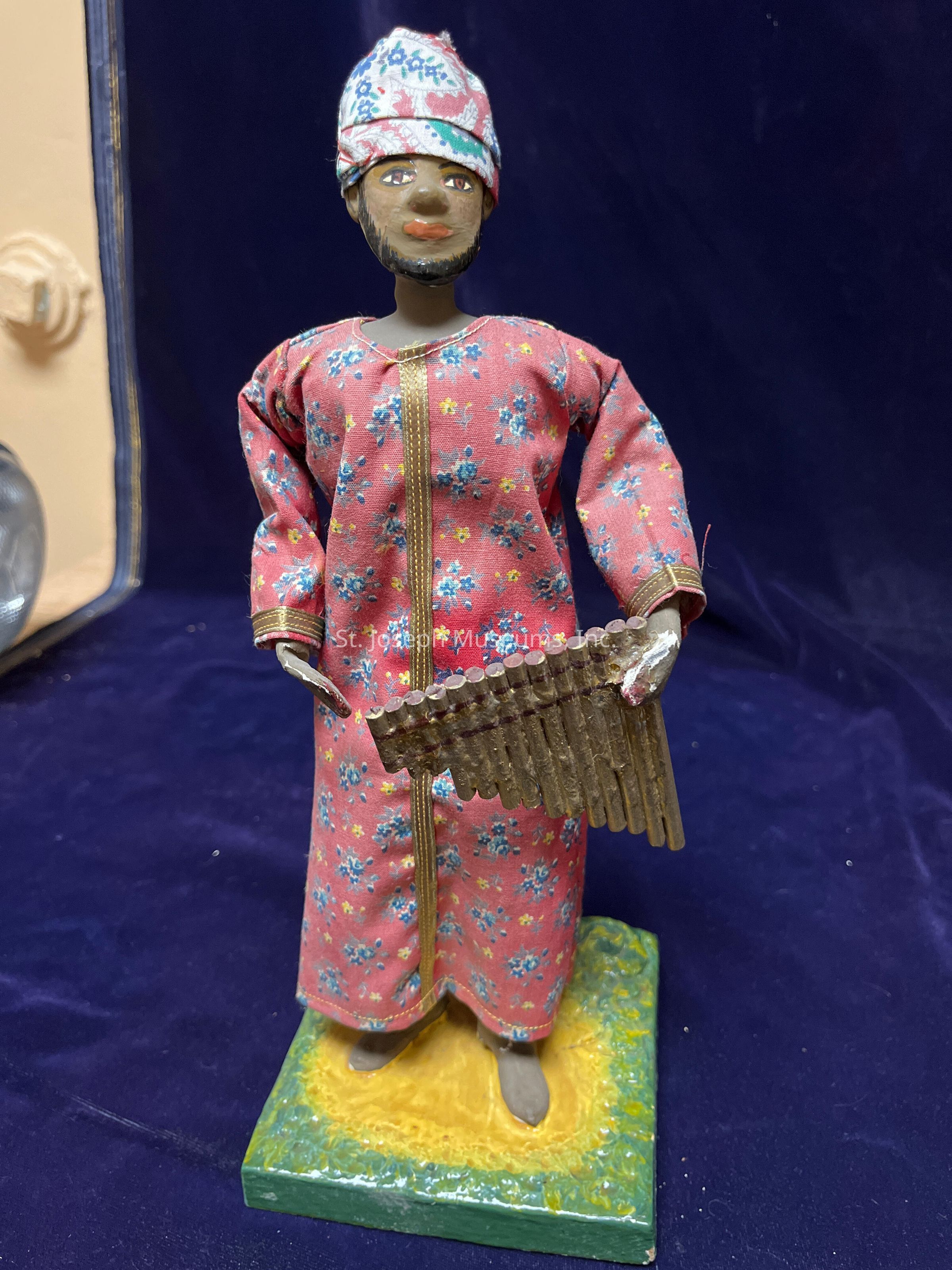 The image depicts a figurine on a textured square base, showcasing a person dressed in a vibrant, floral-patterned outfit with long sleeves, featuring a combination of pink, blue, and yellow hues, accented by golden trim. The figure wears a headwrap with similar floral designs in blue and red, complementing the attire. The facial expression is detailed with painted features, including striking eyes and a mustache, with a small beard enhancing the character's look. The figure holds a traditional musical instrument resembling a pan flute, made of cylindrical pieces, indicating a cultural or artistic representation. The background is a deep blue fabric, drawing focus to the colorful attire and instrument, creating an impression of a unique blend of culture, art, and music in a miniature sculptural form.