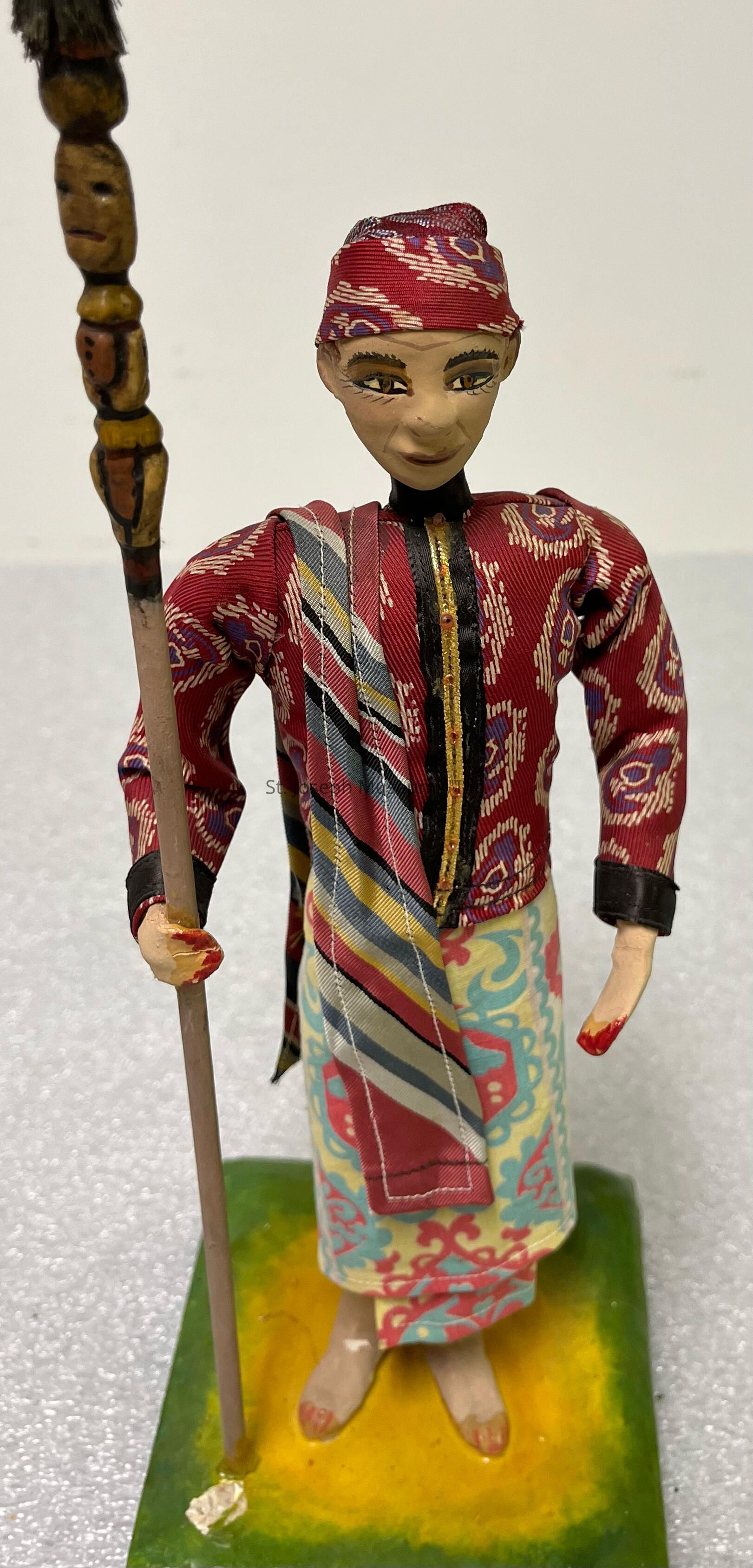 The image showcases a handcrafted figurine of a person dressed in elaborate traditional attire with intricate patterns, featuring a red patterned headscarf and a long tunic made of rich, textured fabric adorned with gold and multicolored designs; the figure holds a tall, carved staff with a face-like carving near the top, adding an air of mysticism to its appearance, while its face displays distinctly painted features, including prominent eyes and a subtle smile, giving it a lifelike expression; the clothing combines various colorful fabrics, including a striped sash draped across the shoulder and a skirt with vibrant blue and red patterns, all standing on a green base, suggesting it is most likely a cultural or ceremonial representation, capturing the essence of a specific ethnic or cultural heritage, with attention to detail in the fabric and painted elements, resulting in a captivating, artistic piece that tells a story through its traditional costume and symbolic accessories.