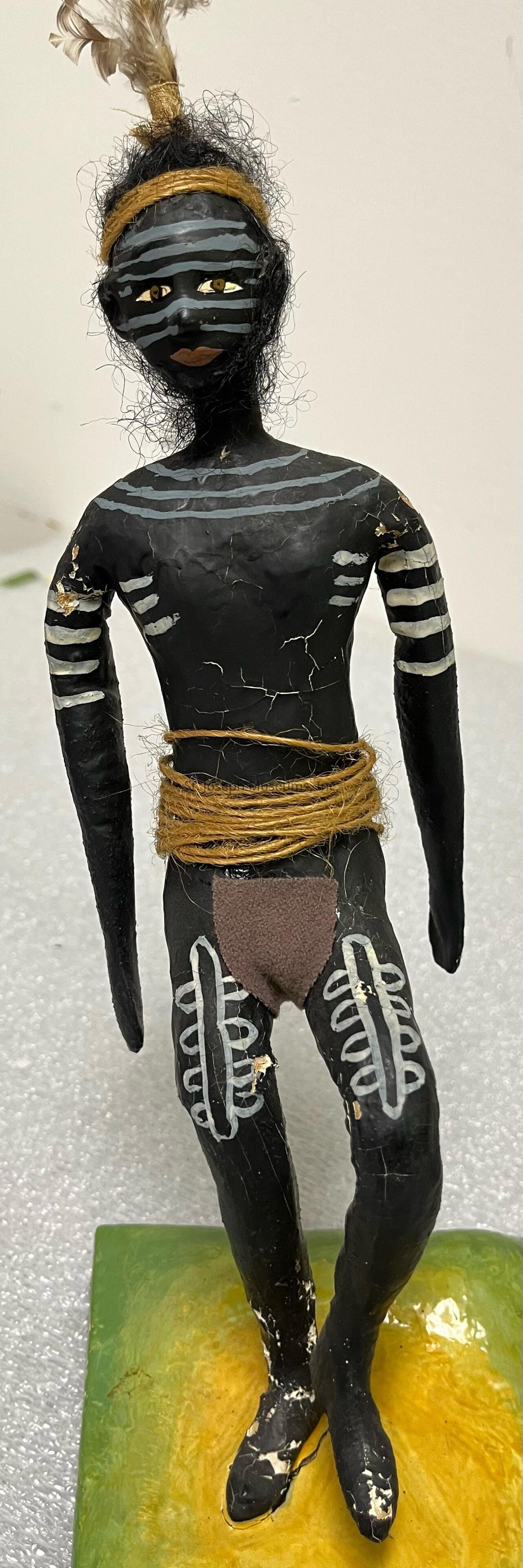 The image depicts an intricately crafted figurine with a striking design. The figure is painted black with white patterns on its face, arms, and legs, giving it a tribal aesthetic. It features a headband wrapped around its head, adorned with feathers, adding a ceremonial touch. The hair is represented by curly black fibers, contributing to its textured look. Wrapped around the waist is a string, accentuating its detailed costume, and a small loincloth covers the lower waist. The figurine stands firmly on a green and yellow base, suggesting a natural setting. Each element combines to form a piece that highlights artistic creativity and cultural inspiration.