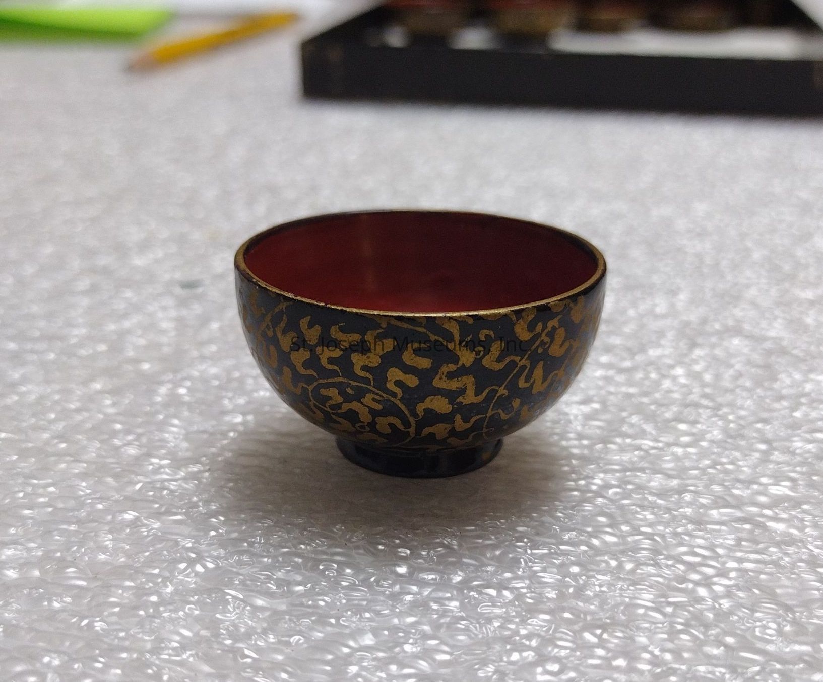 The image showcases a small, intricately designed bowl with a dark exterior adorned with golden, organic patterns resembling vines or tendrils, giving it an elegant and traditional appearance. Inside, the bowl is a rich, deep red, providing a striking contrast that highlights the craftsmanship. The bowl is resting on a textured, bubble-wrap-like surface, which suggests care in handling or storage, possibly indicative of its value or fragility. In the background, there's a slight blur that reveals part of another object, perhaps a pencil and the edge of a dark box, suggesting a work or display area. The composition of the image draws attention to the bowl’s detailed pattern and luxurious finish, emphasizing its aesthetic appeal.