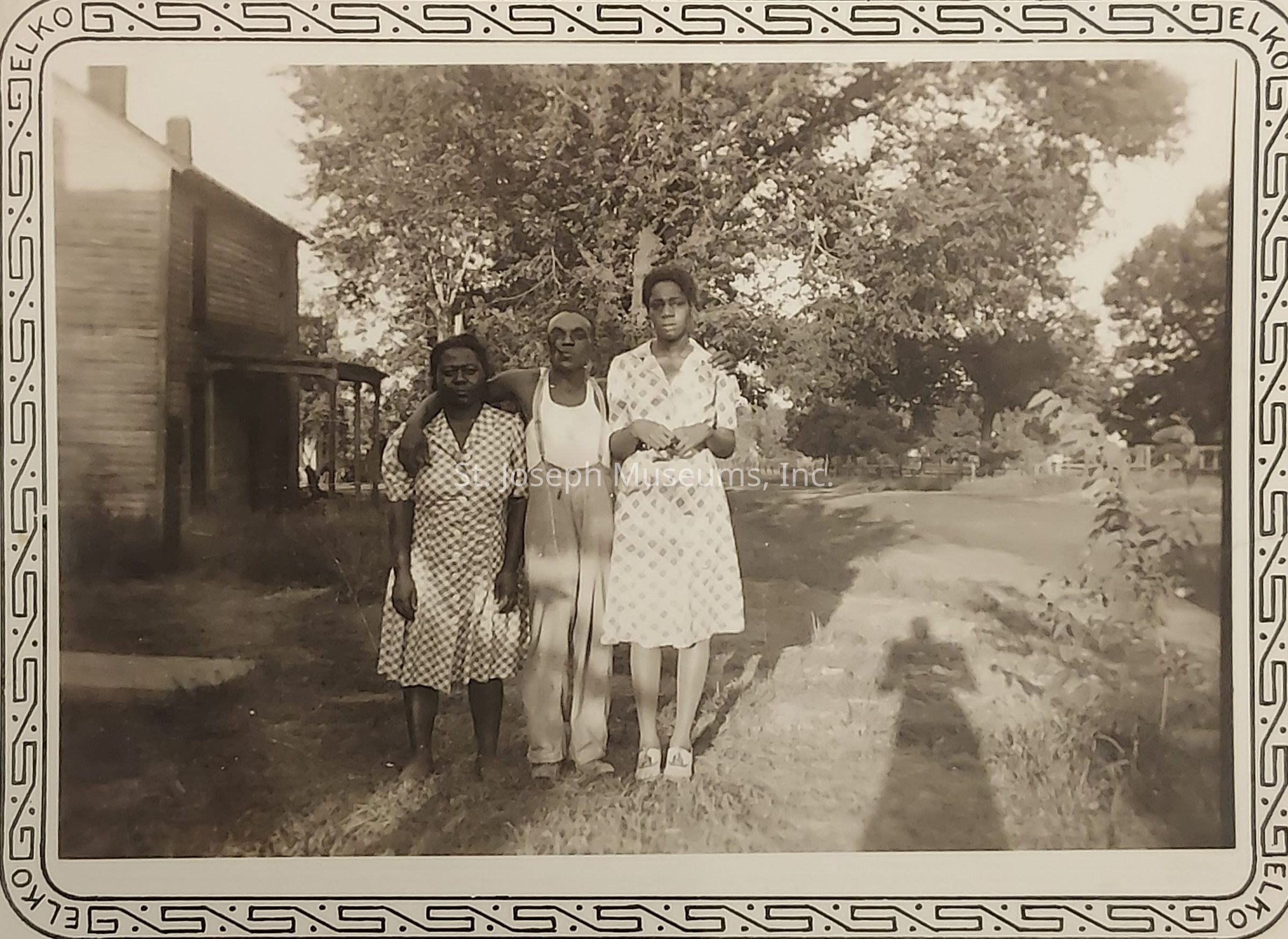 The image depicts three individuals standing closely together outdoors, with two women flanking a man who has his arms affectionately draped around them; they are positioned on a grassy area with a dirt path to the side, and in the background is a wooden building and leafy trees, casting a patterned light and shadow play across the scene. They are dressed in period clothing, possibly from the mid-20th century, with the women wearing patterned dresses and the man in suspenders and a sleeveless top. The border around the photograph adds a decorative touch, and the shadow of the person taking the photo stretches across the grass towards the trio, suggesting it was taken in the late afternoon or early evening, capturing a moment of familial or friendly connection and warmth against a serene and rural backdrop.