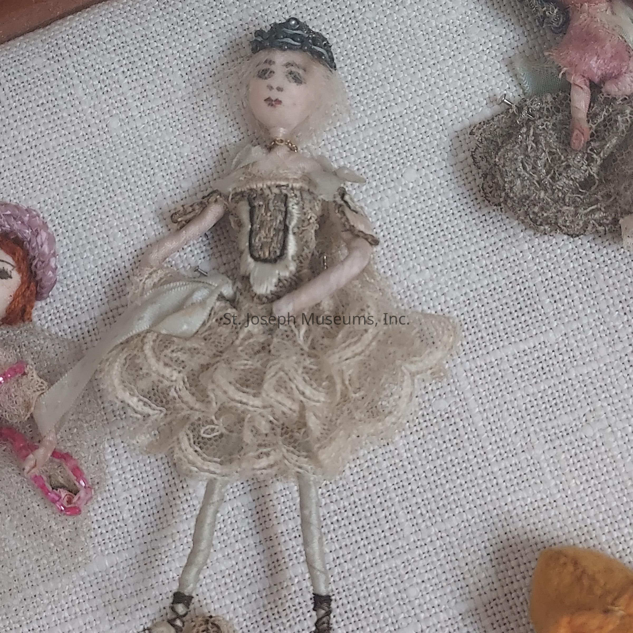 The image features a small, intricately crafted doll with delicate facial features sitting on a textured, light-colored fabric background. The doll is dressed in an ornate costume, showcasing a lace-like, vintage-style dress with layered ruffles and fine details that suggest careful craftsmanship. Its arms and legs, thin and extended, suggest a sense of elegance and poise. The doll also wears a decorative headpiece, adding to its overall antique aesthetic. The detailing on the fabric and the embroidery on its attire are intricate, highlighting the effort and skill involved in creating such a miniature piece. The surrounding objects, although partially visible, include another similarly styled figurine, contributing to a theme of antique or vintage charm. The overall composition and detailed elements capture a sense of nostalgia and meticulous artistry, giving the impression of a cherished collectible or heirloom. The photo showcases not only the beauty of the doll itself but also a broader appreciation for traditional craftsmanship and aesthetic value.
