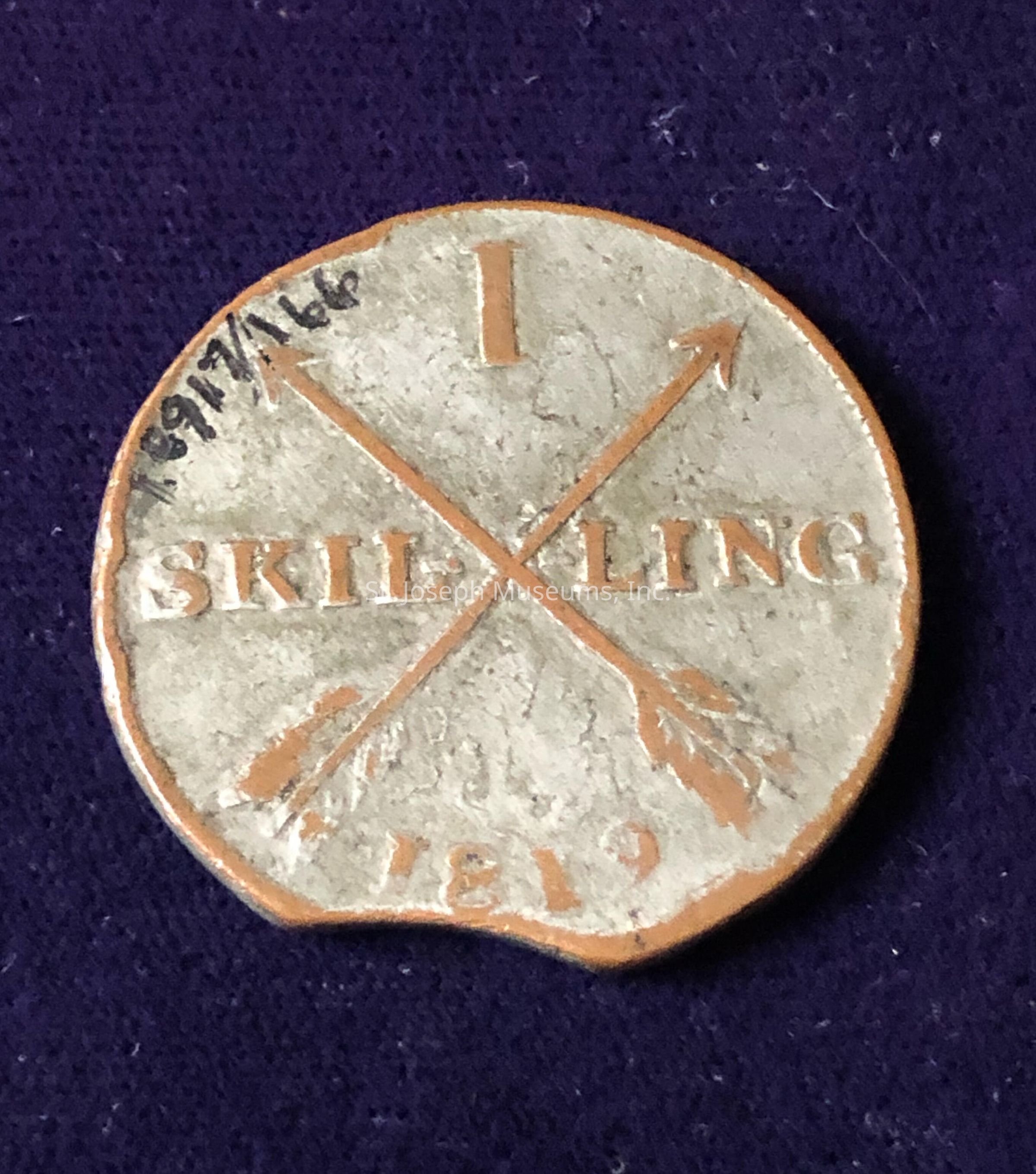 The coin is inscribed with the denomination “1 SKILLING” and dated “1819” at the bottom. The design includes two crossed arrows, adding a decorative element to the surface. The edges of the coin appear worn and slightly tarnished, indicating its age. Some handwritten numbers are visible at the top left, possibly referencing a date or catalog number, written with a fine, dark ink. The overall aesthetic of the coin conveys a sense of historical value and numismatic interest.