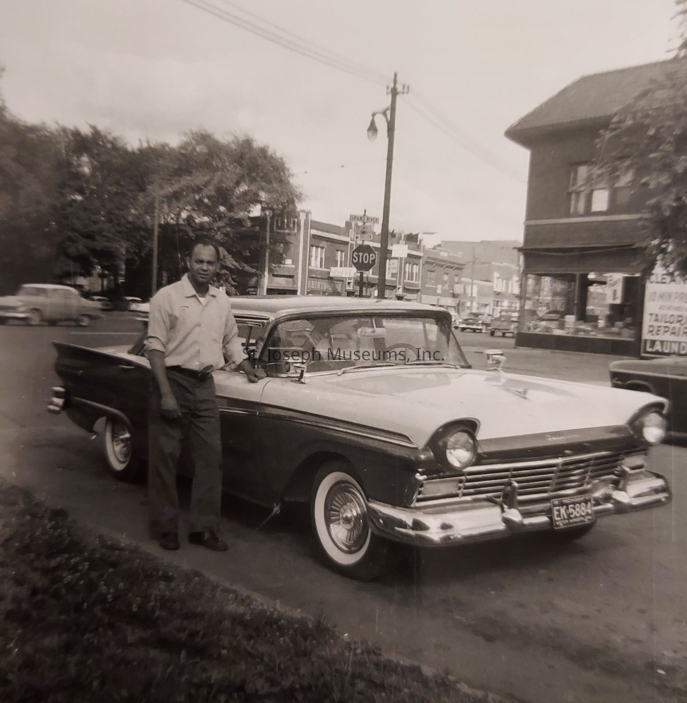 In this vintage black and white image, a man stands confidently beside a classic car, parked along a street in a town that exudes a mid-20th-century charm, characterized by its distinct architectural style and the visible bustling storefronts in the background; the town setting includes a "STOP" sign and various buildings with signs including "TAILOR," "REPAIR," and "LAUNDRY," while other vintage vehicles can be seen driving or parked in the distance, contributing to the nostalgic ambiance of the scene under a partially cloudy sky.