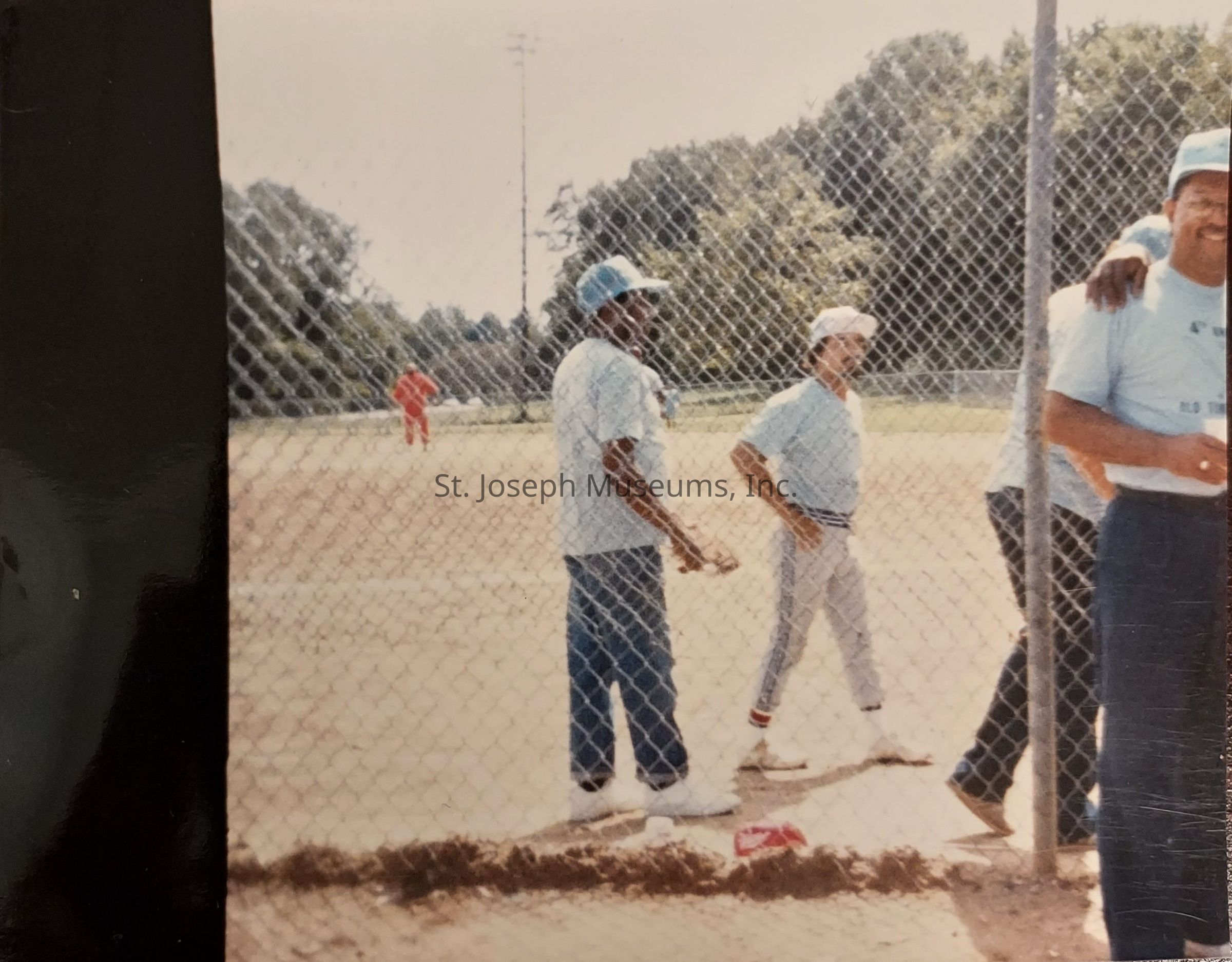 In this vintage photograph, a group of men dressed in light blue shirts, dark pants, and baseball caps are standing near a chain-link fence on a baseball field, with some engaged in conversation, while others casually observe the action on the field; in the background, a person in a red outfit can be seen on the grassy area of the diamond, surrounded by lush green trees, suggesting a warm, sunny day perfect for a friendly game of baseball, as the mood appears relaxed and jovial, capturing a sense of camaraderie and the leisurely enjoyment of sports in a community setting.