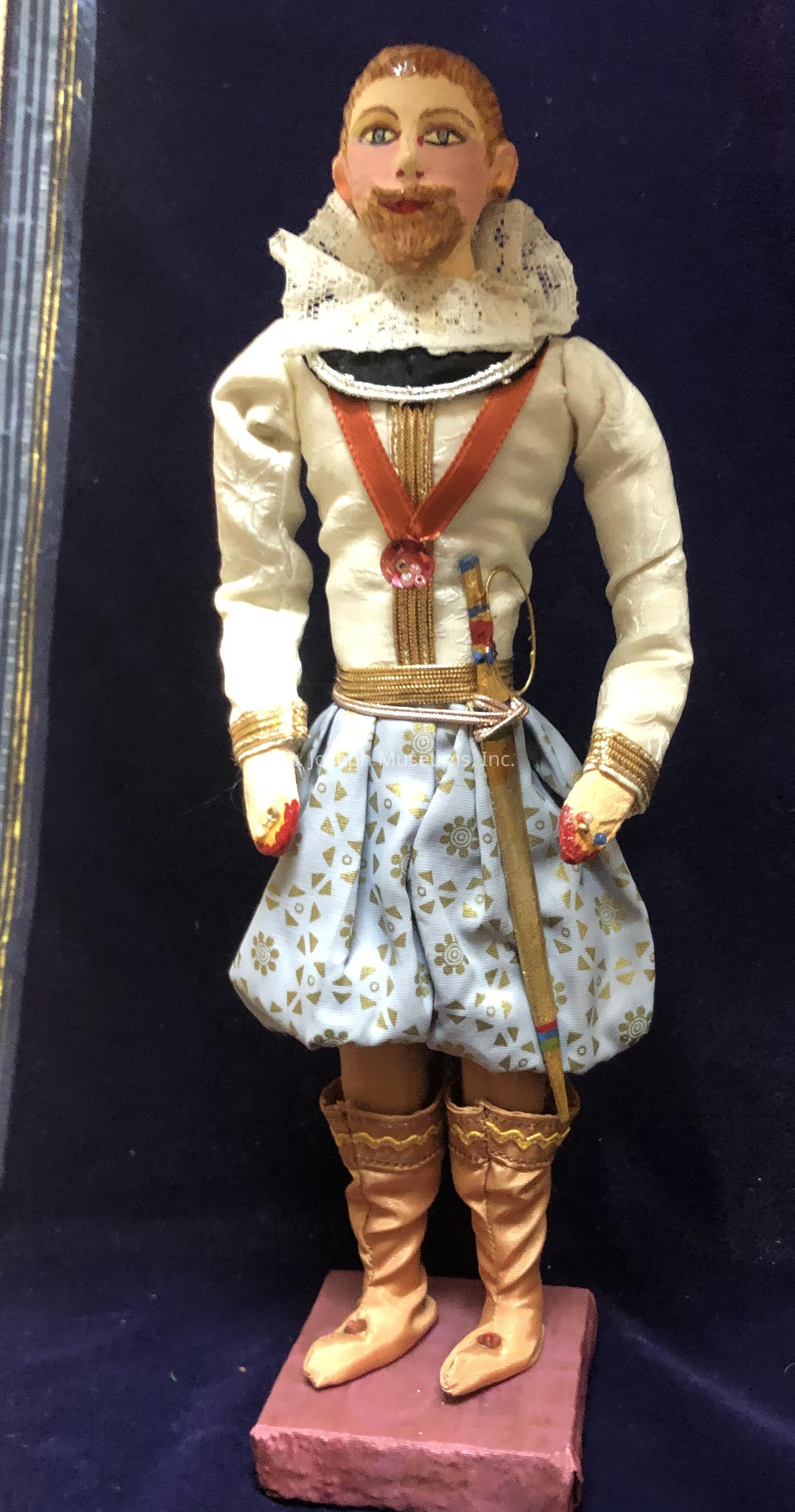 The image shows an intricately designed doll standing against a dark blue background, dressed in a historical or theatrical costume. The doll wears a white lace ruff around its neck, a cream-colored shirt with long sleeves and cuffs adorned with gold trim, and a red sash crossing its torso. The lower half is adorned with light blue, intricately patterned trousers, decorated with small gold geometric shapes and floral motifs. The doll also sports brown boots that reach halfway up its legs, each boot detailed with yellow embellishments at the top. It holds a decorative object that resembles a sword in its belt. The figure has a painted face with strikingly detailed blue eyes, and a small beard, adding a touch of realism and personality to its expressive demeanor. The doll is mounted on a simple, rectangular base, which supports it in an upright position. Overall, its ornate design and historical attire give it an air of regality and charm.