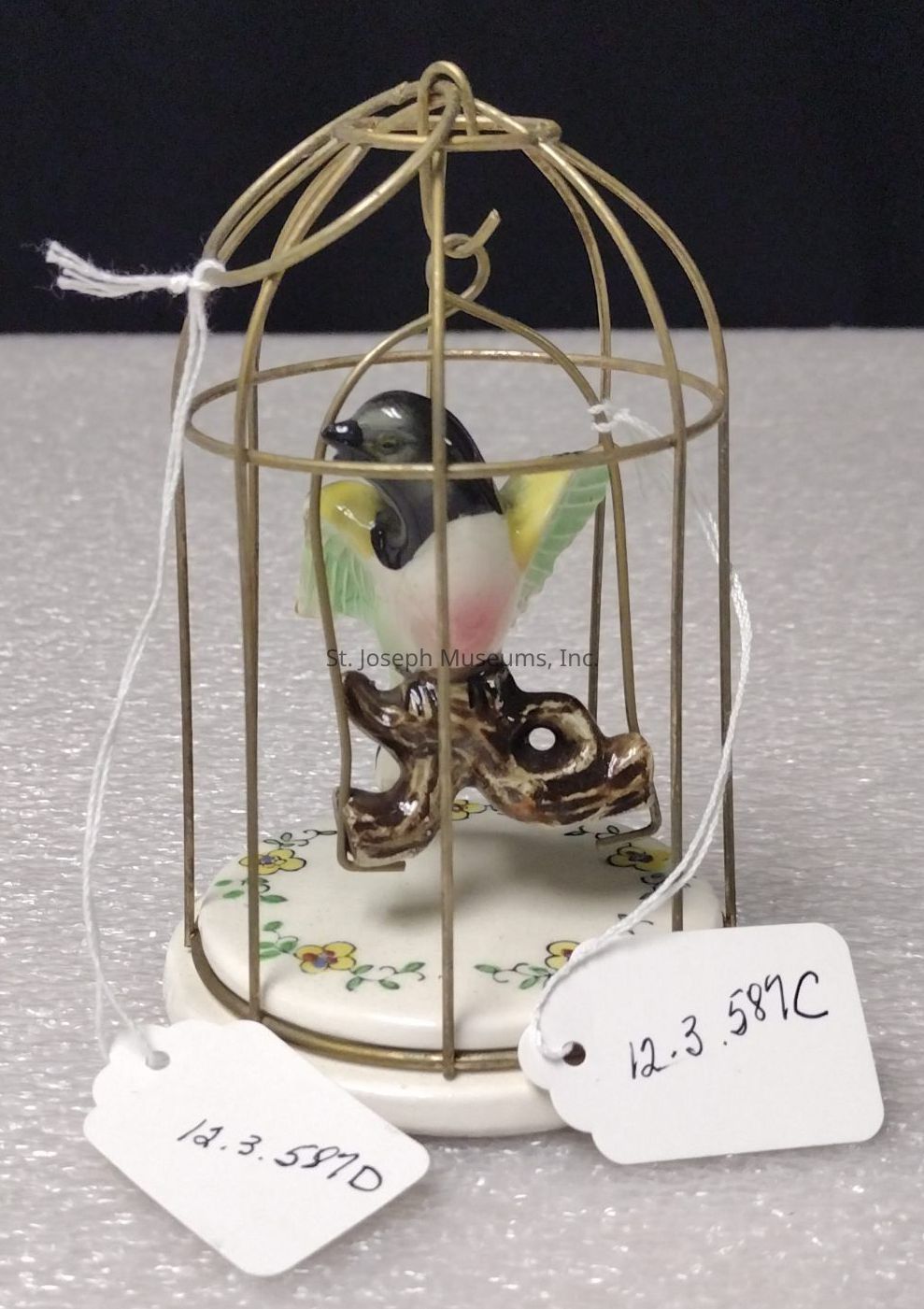 The image depicts a delicate ceramic figurine of a bird with vibrant green, yellow, and pink colors, perched on a small branch inside an intricately designed, gold-colored metal birdcage, which rests on a round porcelain base adorned with hand-painted floral patterns, while two white tags with handwritten labels hang from the cage, presumably providing identification or catalog numbers for the item, and the entire scene is set against a dark backdrop on a light-textured surface, highlighting the ornamental birdcage as a captivating piece of art or decor.