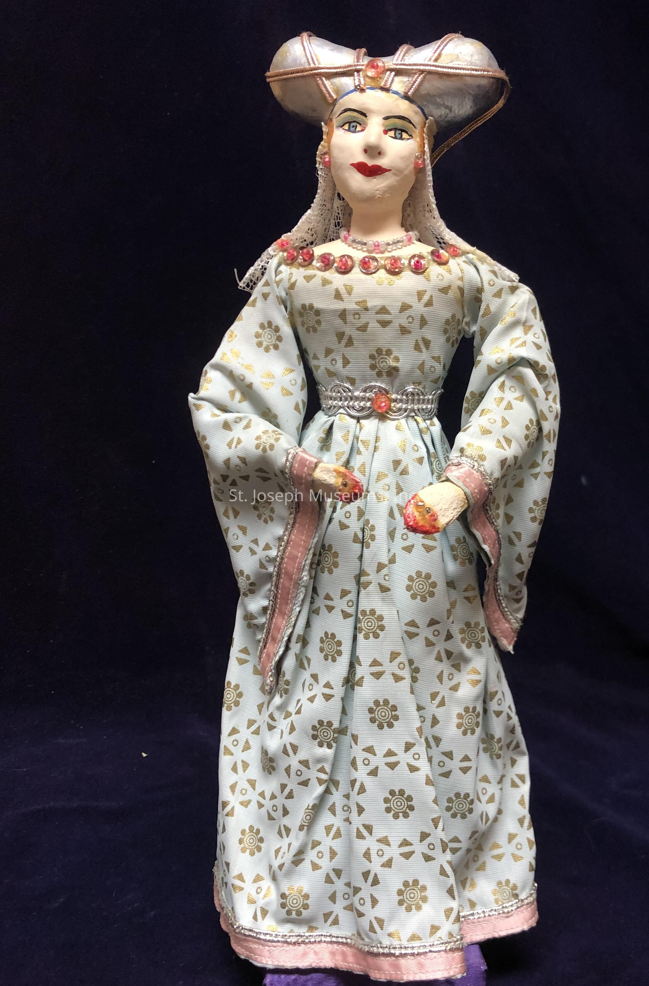 The image showcases a finely crafted doll dressed in an elegant historical costume, featuring a long, patterned fabric in soft colors adorned with intricate gold designs, which is complemented by wide sleeves and a detailed waistline. The doll wears a headdress with a unique, elaborate shape, enhancing its regal and traditional appearance. The facial features are delicately painted, displaying striking eyes and a gentle smile, while the neckline is accentuated with red embellishments, adding a touch of color and sophistication. The doll is set against a dark background, highlighting its intricate details and craftsmanship.