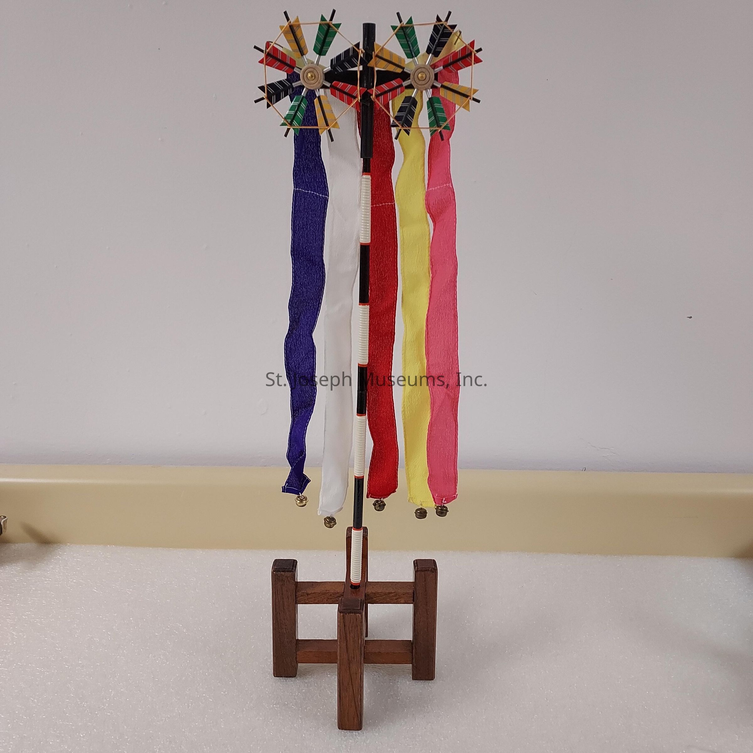 The image displays a decorative object composed of two intricate pinwheel-like structures mounted on a vertical pole, which is secured in a wooden base. Each pinwheel design is symmetrical, featuring wooden pegs arranged in a circular pattern, and adorned with vibrant sections of colors including red, yellow, and green. Extending from the structure are four long fabric ribbons in different colors: purple, white, yellow, and pink, each with a small bell attached at the end, providing an element of movement and sound. The background is a plain, neutral wall, allowing the colorful ribbons and intricate structure to stand out prominently against the subdued tones. This artistic piece is potentially used for decoration or ceremonial purposes and exemplifies skilled craftsmanship in its precise construction and vibrant presentation, capturing attention through its blend of color and texture.