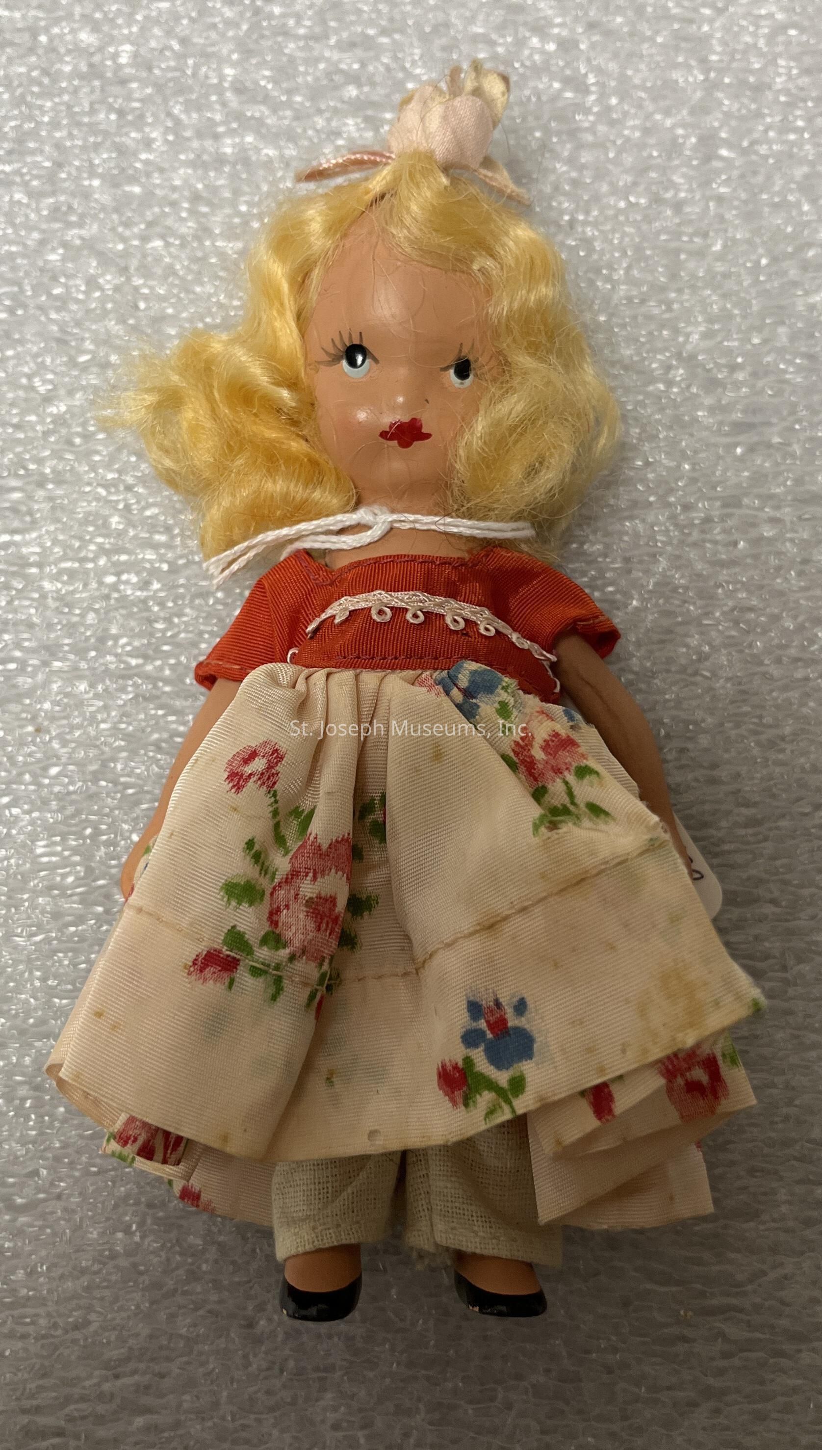 The image features a vintage doll with blonde, curly hair adorned with a small fabric bow on top. The doll has a delicately painted face, highlighted by bright red lips and expressive eyes. She is dressed in a charming outfit consisting of a red top with lace detailing and a floral-patterned skirt that falls over cream-colored trousers. The skirt is decorated with red and blue flowers, adding a quaint and nostalgic touch. The doll's shoes are small and black, complementing her overall appearance. The background is a textured, light material, which contrasts with the vivid colors of the doll's attire, enhancing her presence in the image.