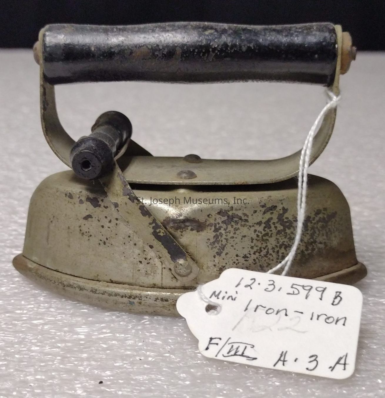 The image depicts an old, vintage iron with a dark wooden or bakelite handle and a tarnished metal body, indicative of its age and frequent use, suggesting it might have been hand-operated rather than electric; there is a white string attached to the handle, from which hangs a tag labeled "1.2.3.599B Mini Iron - Iron," alongside other notations or codes, suggesting it might be part of a museum or antique collection, where historical artifacts are meticulously cataloged. The iron is resting on a textured, possibly plastic surface, which contrasts sharply with the patina and wear of the iron, evoking a sense of the past juxtaposed with the present. The overall condition of the metal exhibits rust and speckling, highlighting the passage of time and use. The design is basic yet functional, likely intended for use in smaller or delicate ironing tasks, such as children's clothing or minor garments. The simplicity and robustness of the construction hint at a time when household items were built