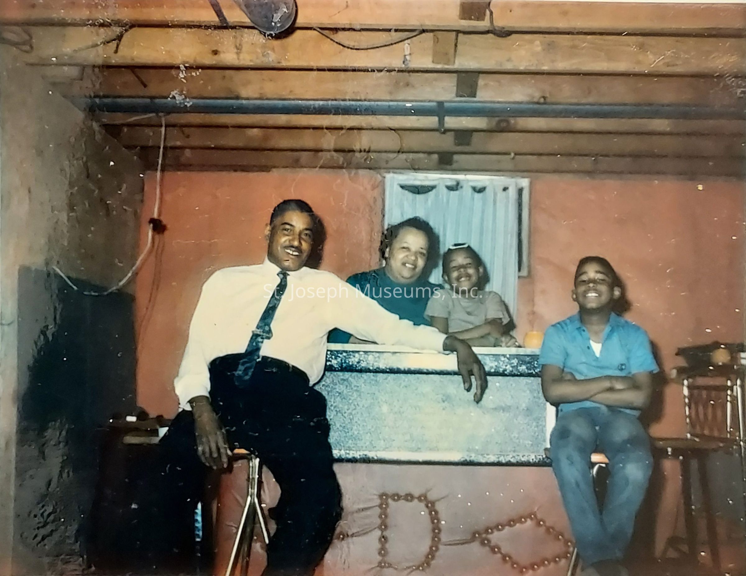 The image depicts a joyful scene in a cozy room, where a family of four is gathered, exuding warmth and happiness. On the left, a man dressed in a white shirt and black tie leans comfortably on a chair, showcasing a cheerful demeanor. Beside him, a woman in a teal top is seated behind a counter or table, holding a smiling child in her arms. The child, wearing a beige top, is captured in a moment of delight, further accentuated by the presence of another child seated on the right, wearing a blue shirt with arms crossed, grinning broadly. The setting appears to be a room with a wooden beam ceiling, painted walls, and an old-fashioned decor style, suggesting a vintage atmosphere. The overall mood is one of familial love and togetherness, as each member shares a genuine smile, highlighting the affectionate bond among them. This intimate portrayal of family life evokes a sense of nostalgia and captures a timeless moment of contentment and unity.