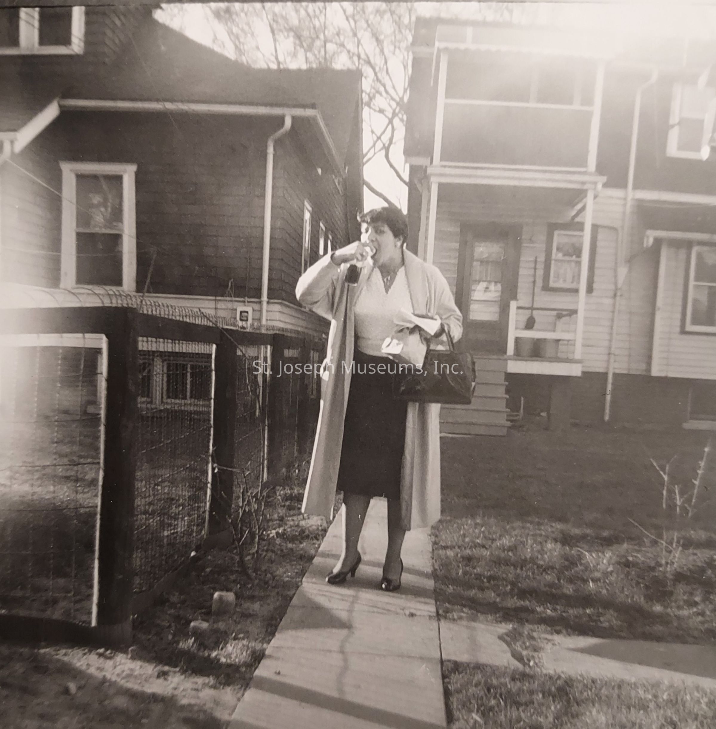In this black-and-white photograph, a woman is captured walking on a narrow path between two houses, wearing a long coat over a white top and a dark skirt, with her feet in pointed shoes. She holds a bag in one hand and is drinking from a bottle with the other. The image appears to be from a previous era, suggested by the style of her clothing and the architecture of the houses. The houses have distinct features; one is partially visible on the left with shingle siding and the other on the right has horizontal siding with steps leading to a front porch. The yard is simple, bordered by a low wire fence on the left, and the trees in the background are bare, indicating it might be during the colder seasons. The woman's stance and the environment around her convey a sense of everyday routine or a moment caught candidly during her daily activities, reflecting a slice of life from that time period.