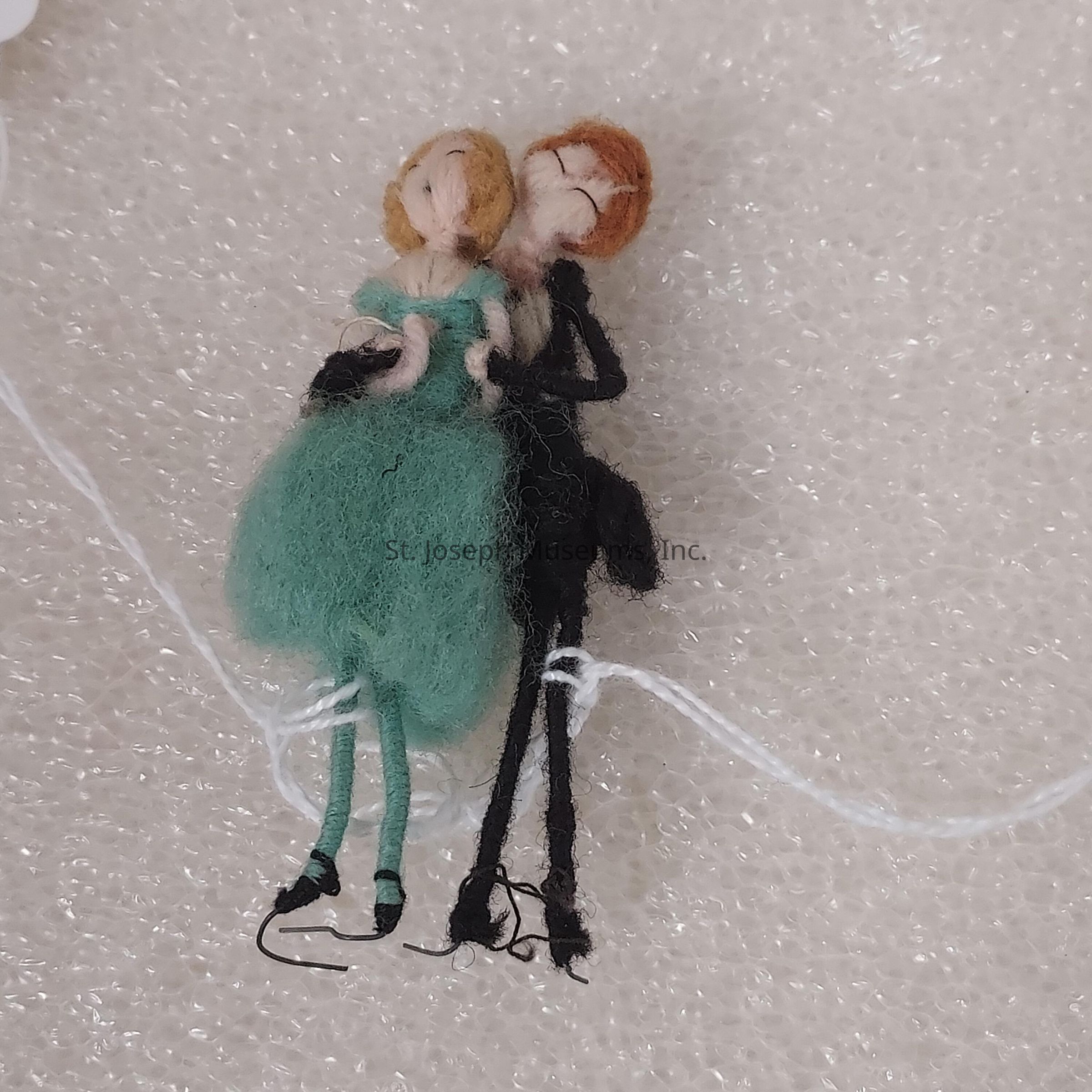 The image depicts two small, intricately crafted felt dolls with delicate features, positioned on a textured, bubbly surface. The dolls are styled as a couple with the left figure, dressed in a green outfit and sporting light hair, and the right figure, adorned in darker attire with reddish-brown hair. The dolls appear to be holding each other affectionately, with detailing in their design emphasizing simple, yet expressive postures such as a loving embrace. The background consists of a white, foam-like material that has a series of small, regular bubbles, providing a soft contrast to the vibrant felt figures. Thin threads, likely a part of the creation process, are visible, adding a handmade, artistic charm to the scene.