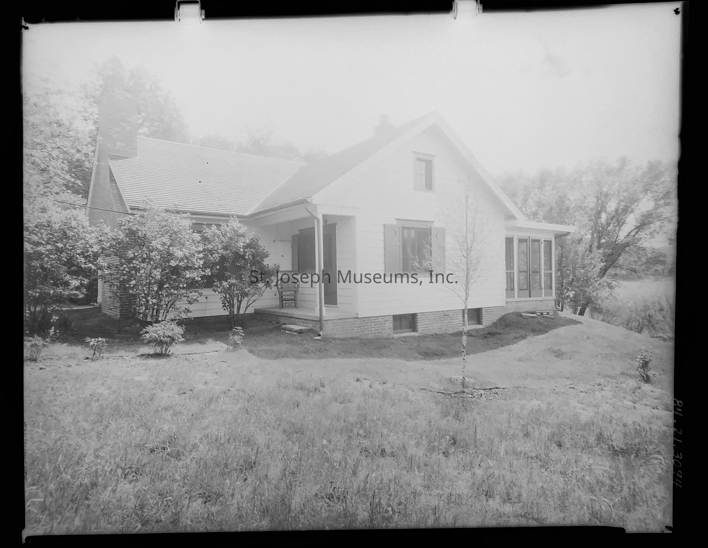 A black-and-white image depicting the backyard of the original house at 2827 Lovers Lane that once belonged to Robert Hastings in St. Joseph, Missouri. The home features a charming, single-story design with a spacious covered front porch, complete with chairs and a table, a gabled roof, and a prominent brick chimney, all set against a manicured lawn and surrounded by shrubbery.
