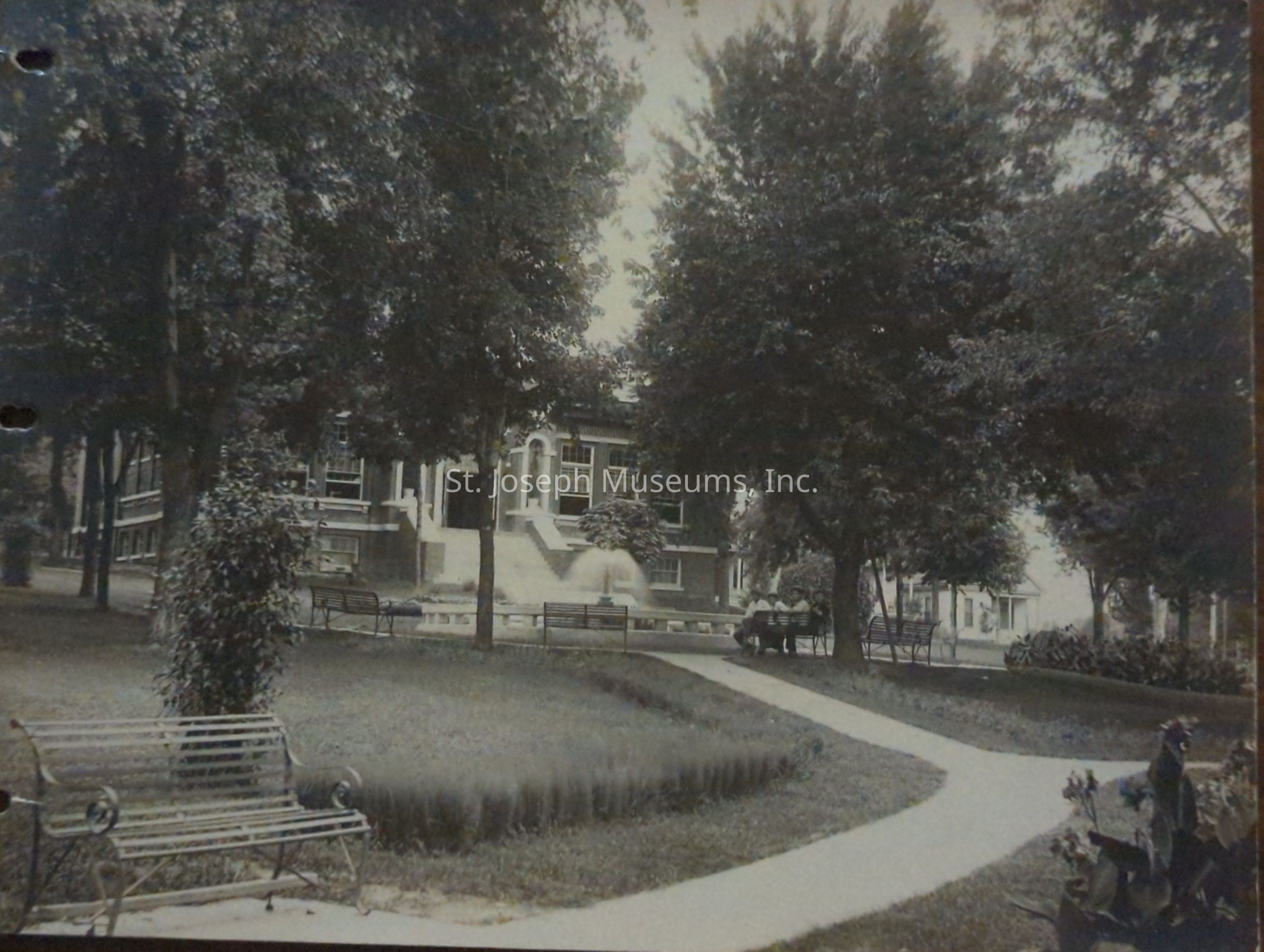 Black-and-white image depicting a serene park setting in front of the Washington Park Library at 1821 North 3rd Street, featuring a pathway curving through well-maintained lawns, several benches, and large trees providing ample shade. The historical library building stands prominently in the background.