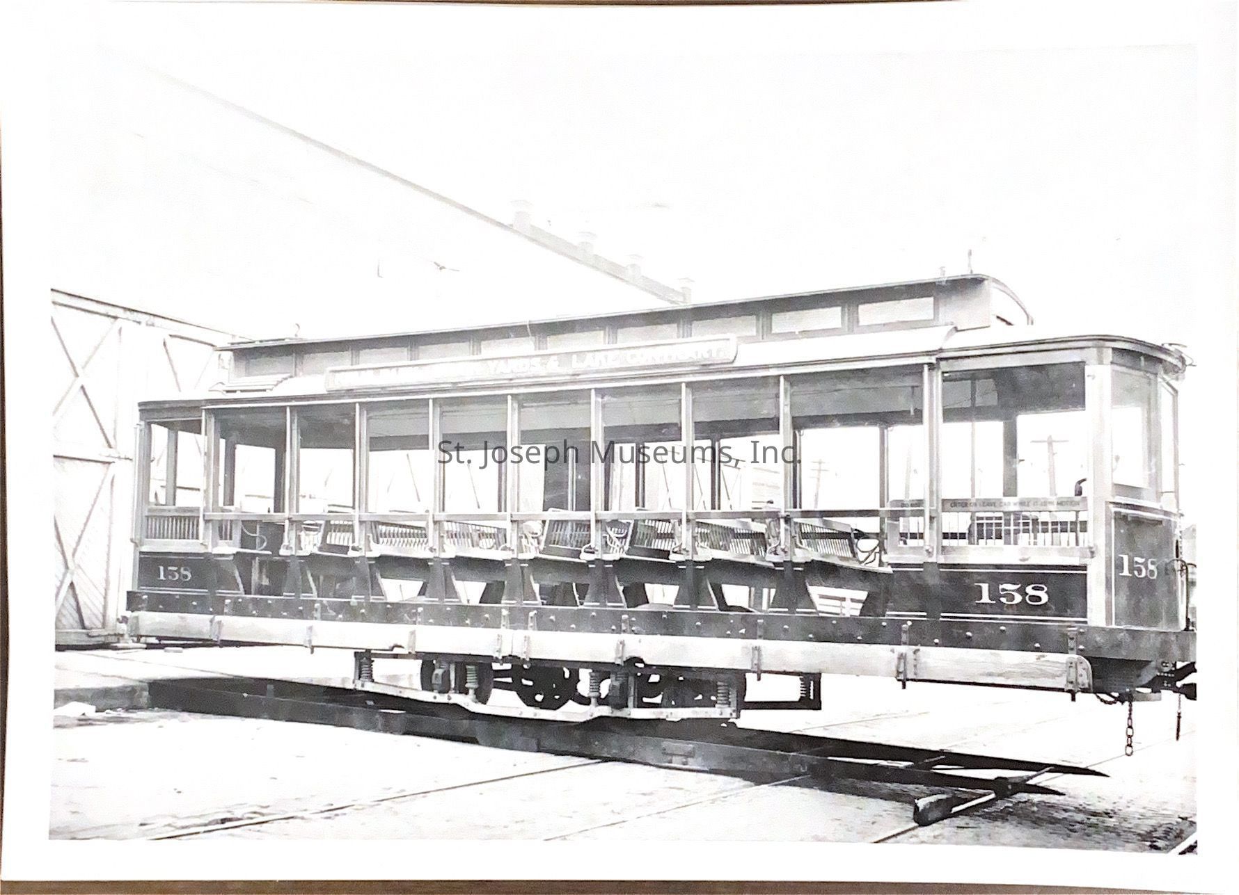 Black and white photograph depicting a vintage streetcar, identified by the number 158 on its side, standing prominently on a track positioned in an industrial setting, likely within a railyard or maintenance area, as it appears to be a photograph from the early 20th century. The streetcar, with its open-air windows and metal framework, features intricate wooden details and sturdy-looking wheels, highlighted by the soft lighting and the backdrop of warehouse-like buildings.